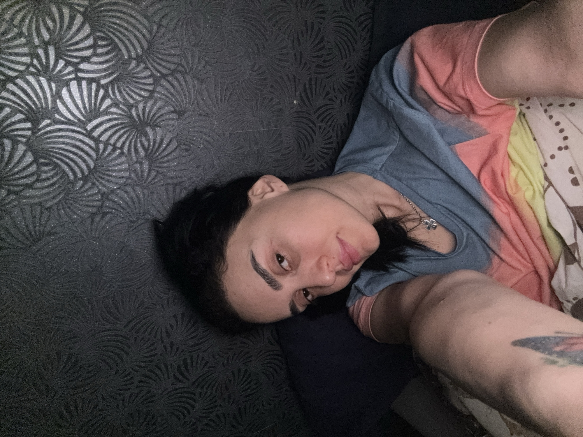 A person with dark hair is lying down on a black patterned surface. They are wearing a blue and pink t-shirt and a silver necklace with a cross pendant. A portion of their right arm is visible, featuring a tattoo. The background is the patterned surface. The location is unknown and cannot be determined from the image.