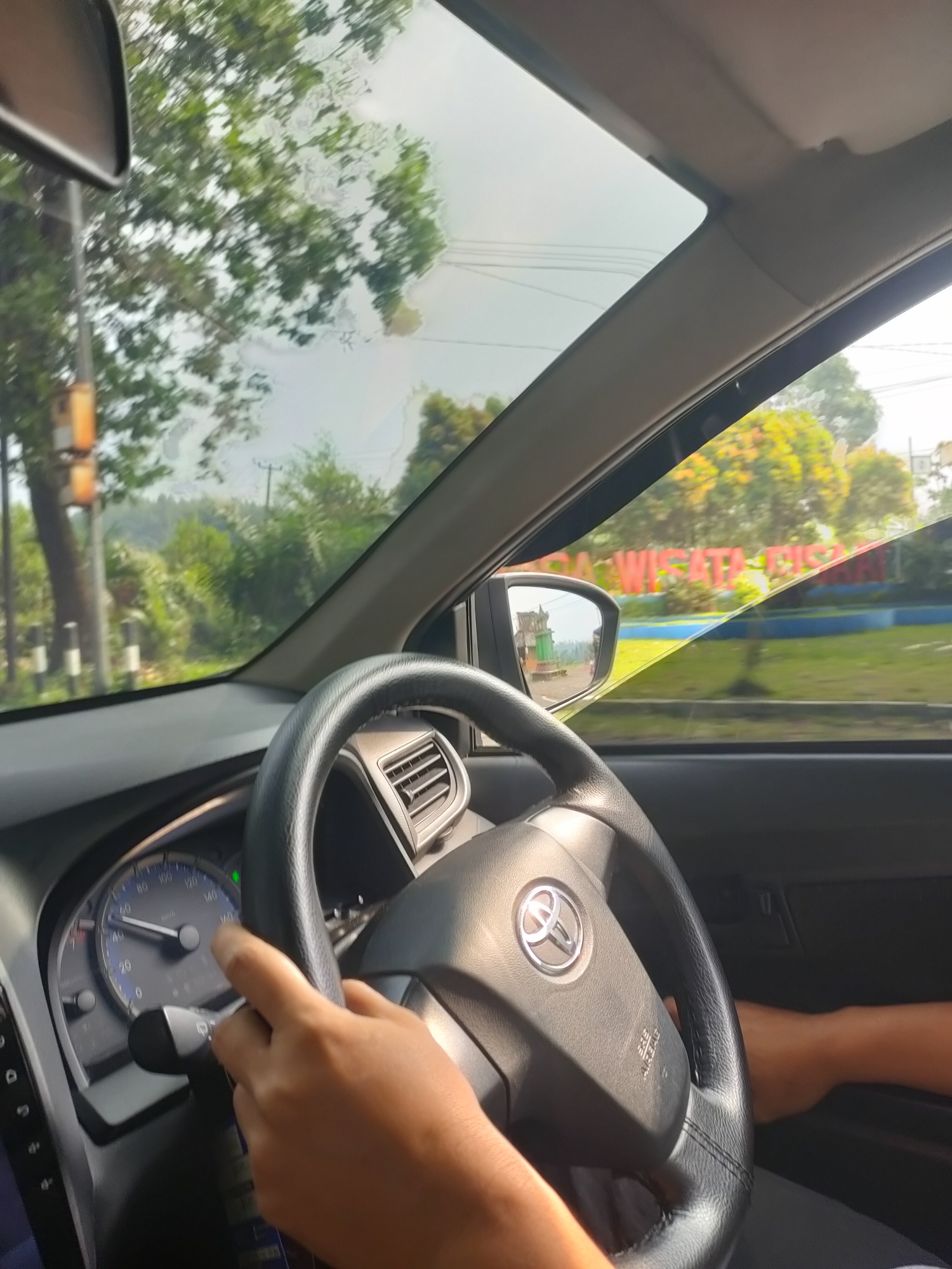 The image is a point-of-view shot from inside a car, likely a Toyota, as it travels along a road in Subang, Indonesia. The driver's hands are visible on the steering wheel and the turn signal/wiper stalk. The dashboard displays the speedometer, indicating the car is in motion. Through the windshield and side windows, lush green trees and vegetation are visible, suggesting a rural or suburban setting. A sign with Indonesian text, possibly related to tourism ("WISATA"), can be seen in the distance. The overall impression is a sunny day and a scenic drive.