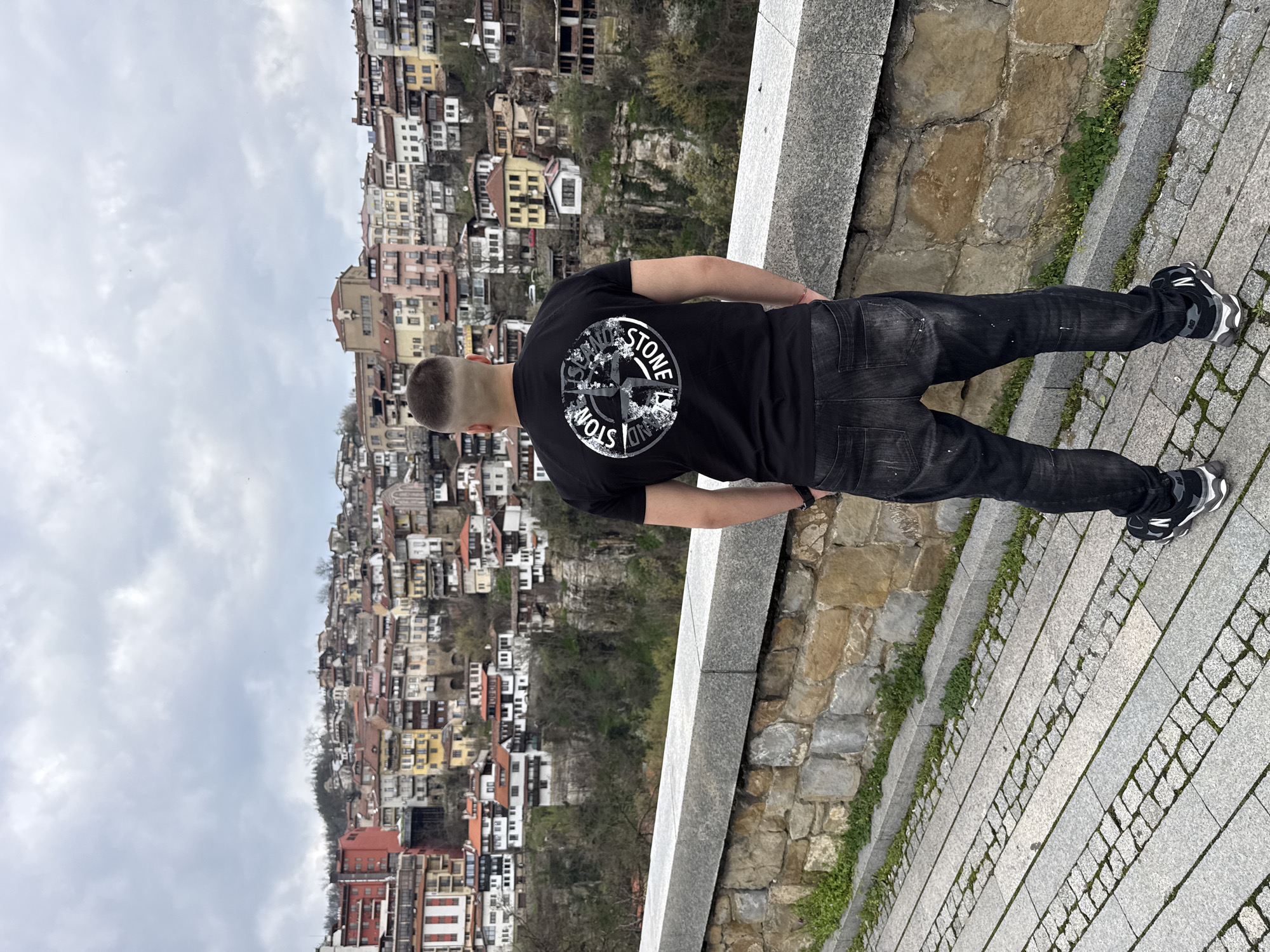 A man stands on a grey stone walkway, facing away from the camera and towards a dense hillside of buildings. The man has a short haircut and wears a black t-shirt with a large, circular graphic on the back, dark jeans with faded areas, and black and grey athletic shoes. He stands with his hands behind his back. To his right, a stone wall forms part of the walkway, with patches of grass growing along its edge. Beyond the walkway and wall, the hillside is densely populated with buildings of varying colors and sizes, appearing as if stacked upon each other. The sky is overcast with a grey, cloudy texture. The location is in the city of Unknown, Unknown.