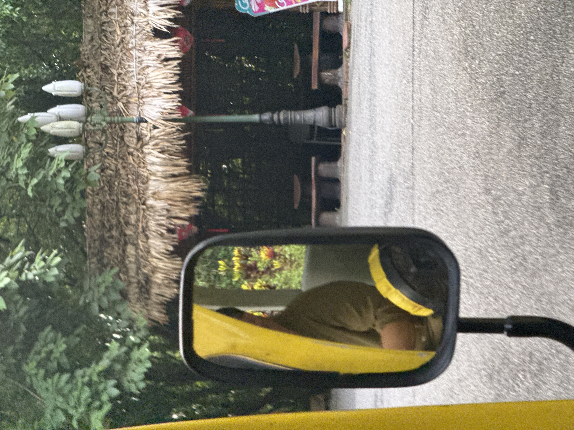 The image shows a side-view mirror of a vehicle, reflecting a person wearing a light brown shirt. The mirror is framed in black plastic and is mounted on a black arm. To the left of the mirror is a bright yellow object, possibly part of the vehicle. In the background, visible through the mirror and to the right, is a thatched-roof structure with a lamp post in front of it. The structure appears to be an open-air pavilion or shelter with stone benches. Red decorative elements hang from the roof. Beyond the structure, lush green foliage suggests a natural or park-like environment. The ground in the foreground is paved with asphalt. The overall impression is that the photograph was taken from within a vehicle moving through a scenic area, possibly a tourist attraction in Tây Hoa Lư Ward, Vietnam, given the architectural style of the shelter.
