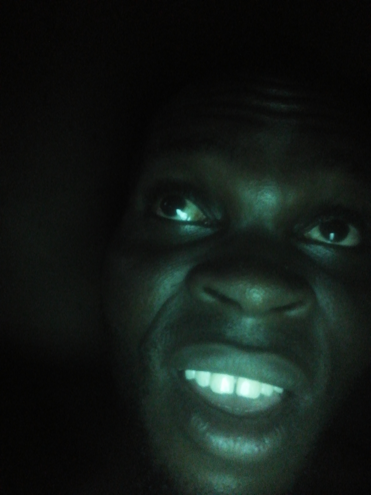 The image depicts a close-up of a dark-skinned person's face, illuminated by a green light. The individual is smiling, revealing their teeth. The background is dark and indistinct. The location is unknown.
