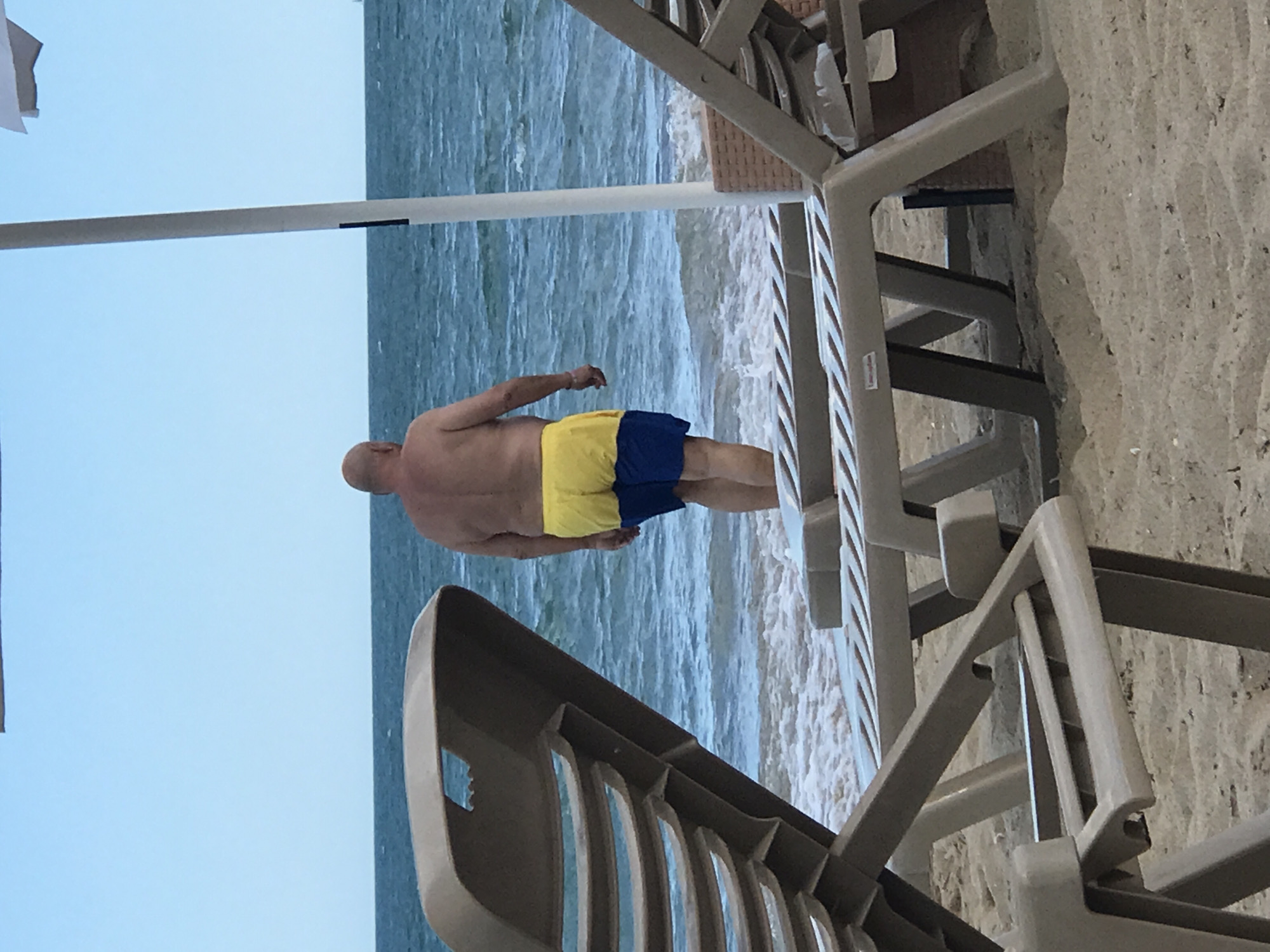 An adult male, appearing to be of middle age and with a bald head, stands facing away from the camera at the edge of the sea on a sandy beach. He is wearing yellow and blue swim trunks. The water is a calm blue with small waves lapping at the shore. In the foreground, two empty, light brown plastic lounge chairs are partially visible. The scene is set on a beach, likely in Varna, Bulgaria, given the context of the image source. The sky is a clear, pale blue.