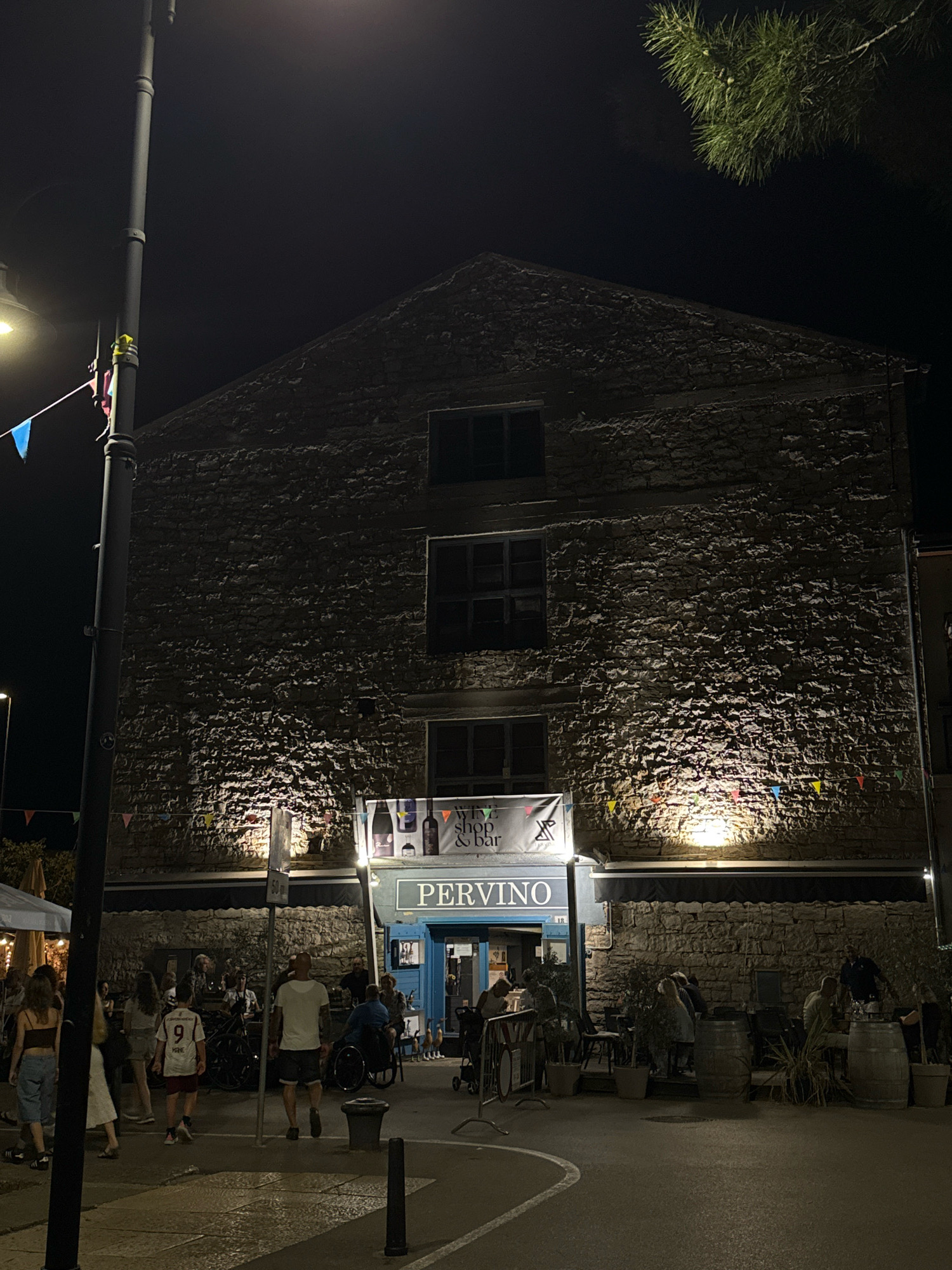 The image depicts a nighttime scene in the city of Grad Novigrad, Croatia, outside a establishment named PERVINO. The building's facade is constructed of rough-hewn stone, with multiple windows visible on its upper floors. A banner above the entrance advertises "Wine Shop & Bar."
In front of PERVINO, people are gathered, some sitting at tables and others standing. The atmosphere appears lively, with string lights and small triangular flags adorning the building. A street lamp illuminates part of the scene, casting long shadows. A person in a wheelchair is visible among the patrons, and several bicycles are parked nearby. The ground in the foreground is paved, with a white line marking a path or road. The overall setting suggests an outdoor dining or social area of a restaurant or bar.