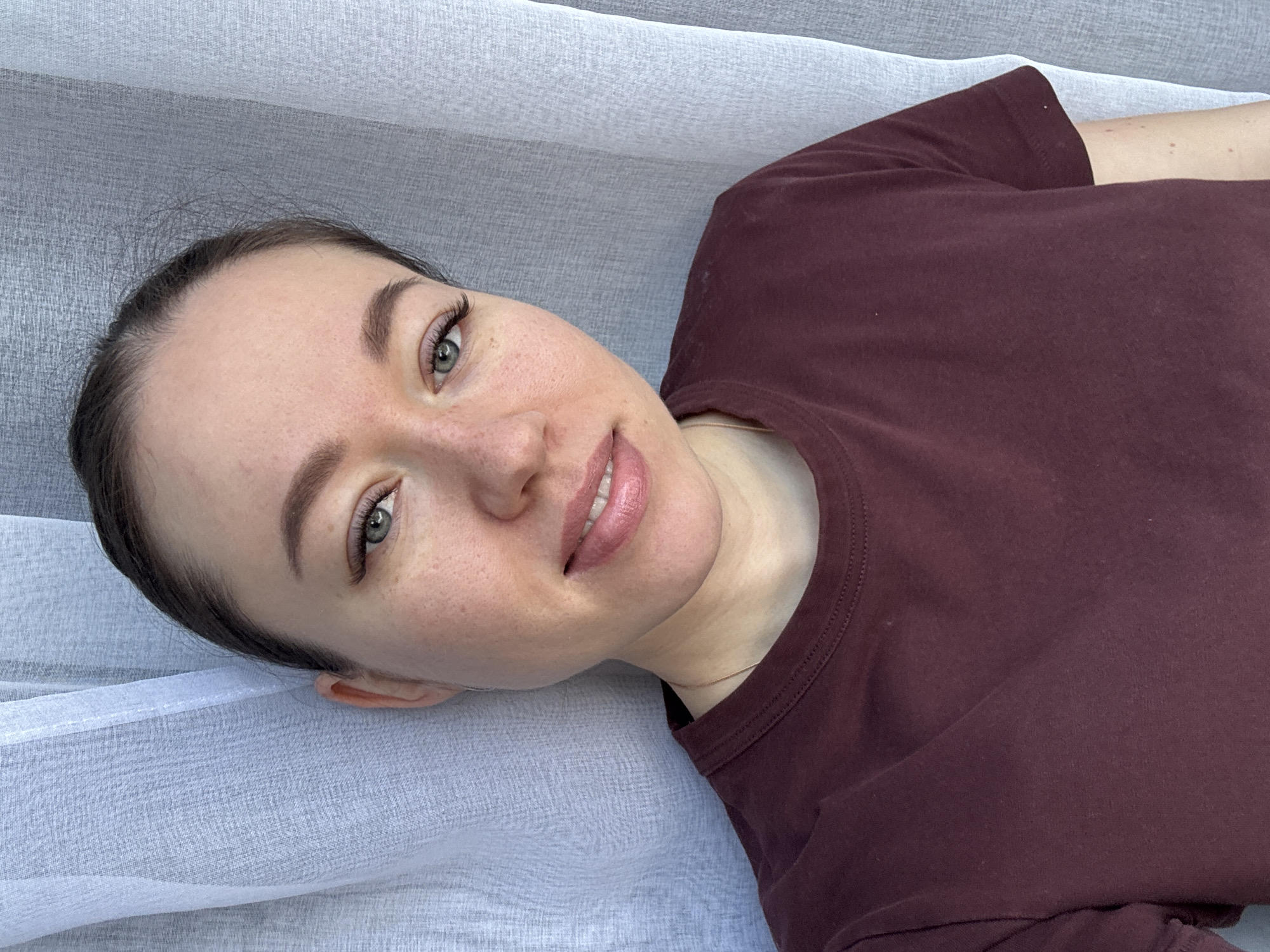 A fair-skinned woman with brown hair, wearing a maroon t-shirt, is lying on a surface covered with a thin, white fabric. Her head is slightly tilted to the left. The location is indeterminate and there is no indication it is in Ferma, Russia.