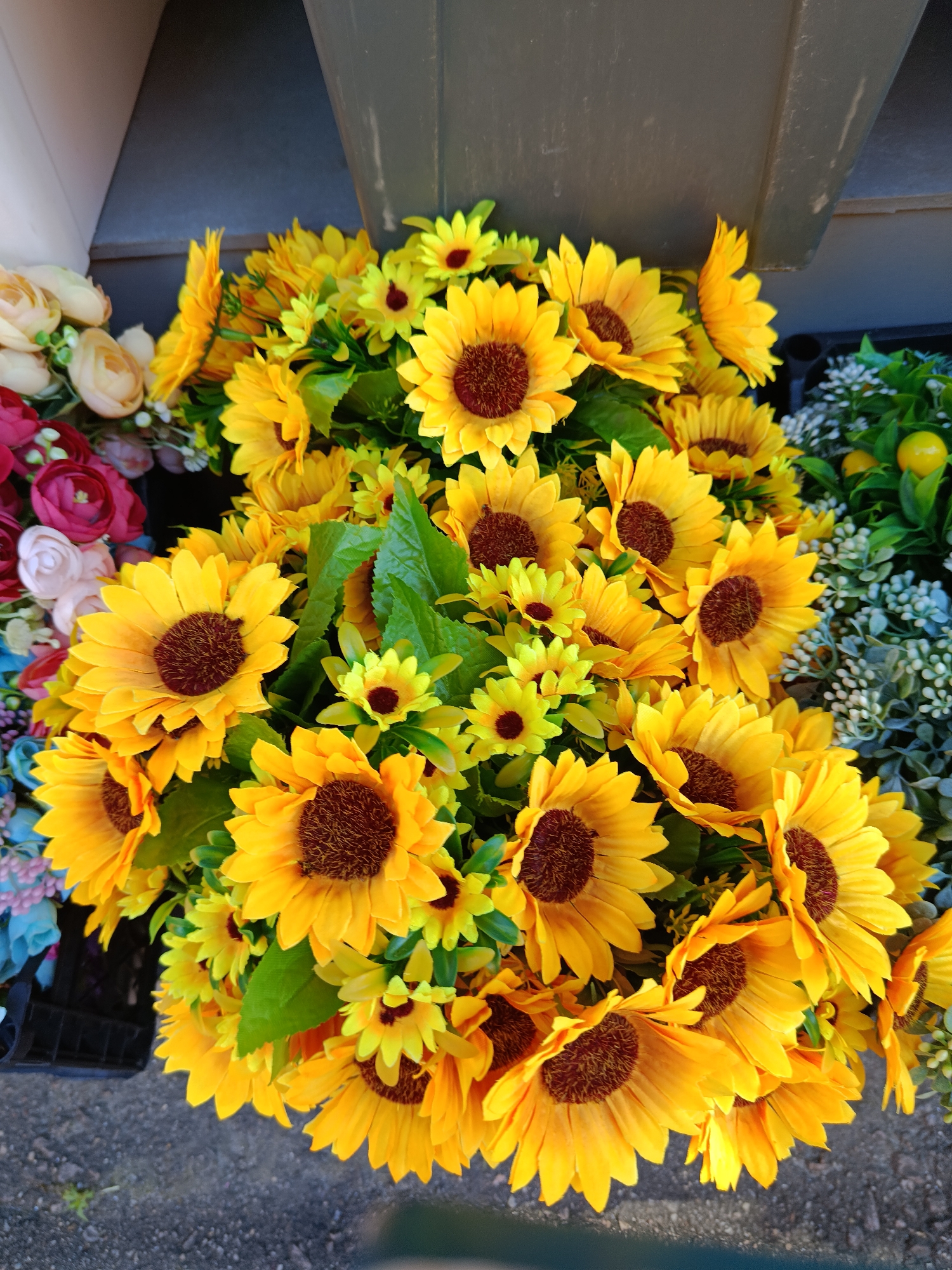 The image shows a close-up, overhead view of a large bouquet of artificial sunflowers and other yellow flowers. The sunflowers are the most prominent, with their bright yellow petals and dark brown centers. They are arranged in a dense cluster, with some smaller yellow flowers interspersed among them. To the left of the sunflowers, there are some artificial roses in shades of pink and red, along with some light pink and cream-colored flowers. In the background, to the right, there is a display of greenery with small white flowers and some artificial yellow fruits, possibly lemons. The flowers appear to be arranged in a planter or bin, with the edge of a dark container visible at the bottom left. The ground beneath the flowers appears to be asphalt or gravel. The scene is well-lit, suggesting it is outdoors or in a brightly lit space. There are no people or animals visible in the image. The overall impression is one of a colorful and vibrant floral display, likely for sale or decoration.