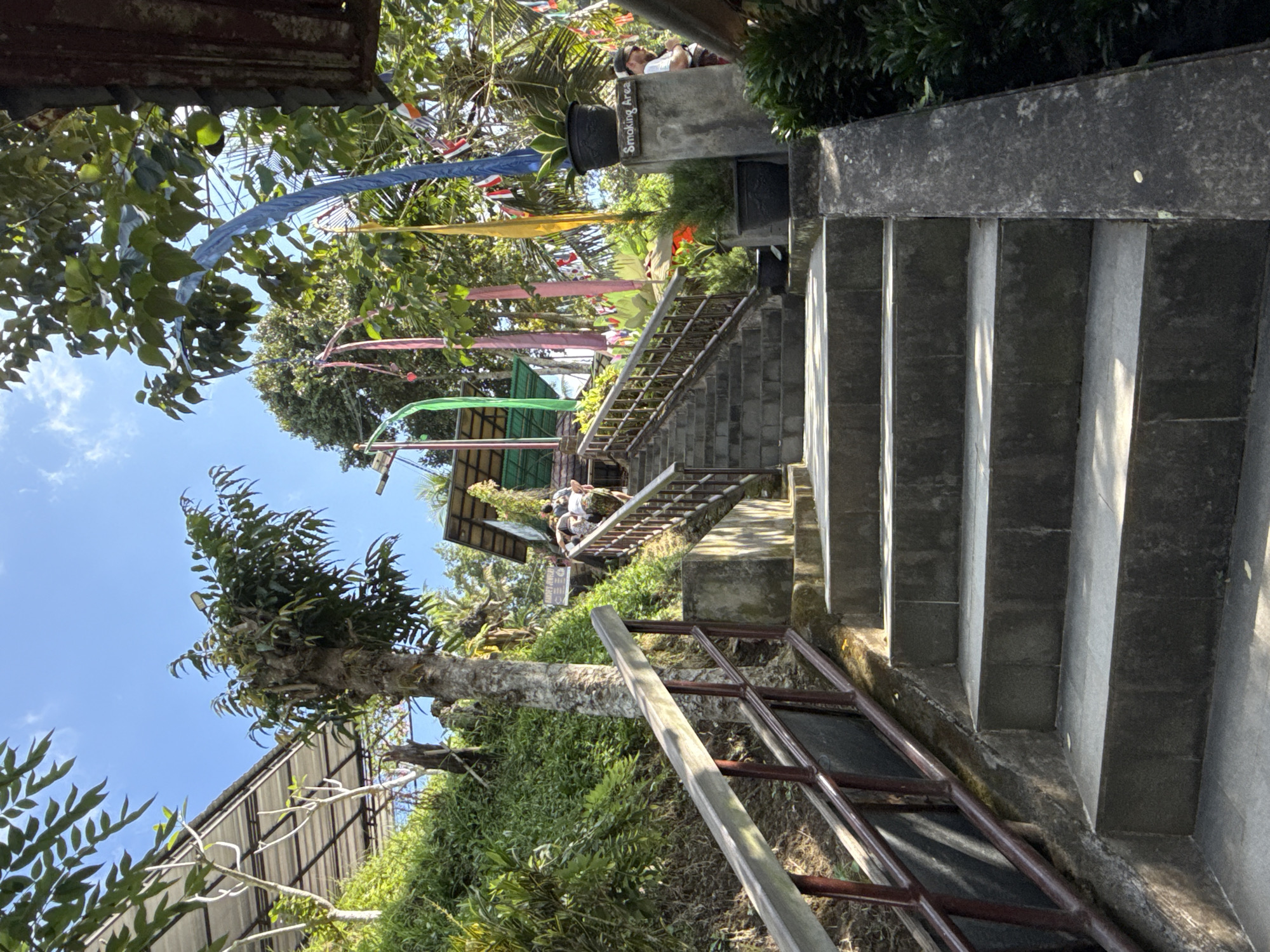 In Tampaksiring, Indonesia, a stone staircase ascends a lush, verdant hillside under a bright blue sky. The stairs are bordered by a sturdy wooden and metal railing on the left and a concrete embankment on the right, which is adorned with potted plants. At the top of the stairs, a building with a green roof is visible, and colorful flags, including yellow, blue, and pink, flutter in the breeze, adding a festive atmosphere to the scene. Several individuals are seen walking up the stairs and gathered near the building. The area appears to be a tourist attraction or a place of worship, with its natural beauty enhanced by decorative elements.