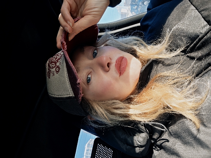 A female individual with blonde, wavy hair and blue eyes is depicted in a medium-close-up, seated inside a vehicle. She is wearing a dark-colored jacket or hoodie and reddish-pink lipstick. A baseball cap is worn on her head, featuring a greyish-beige front panel with a dark reddish emblem and a distressed reddish-burgundy brim. Her right hand is visible, adjusting the brim of the cap. Strong sunlight illuminates the scene from the right, casting distinct shadows on her face and clothing. Through the vehicle's left window, a modern, light-colored, multi-story building with a grid-like facade is visible against a clear blue sky. This contemporary architectural style is characteristic of newer urban development zones within Saint Petersburg, Russia, such as areas in the Primorsky District or the western part of Vasilievsky Island, as opposed to the historic central districts.