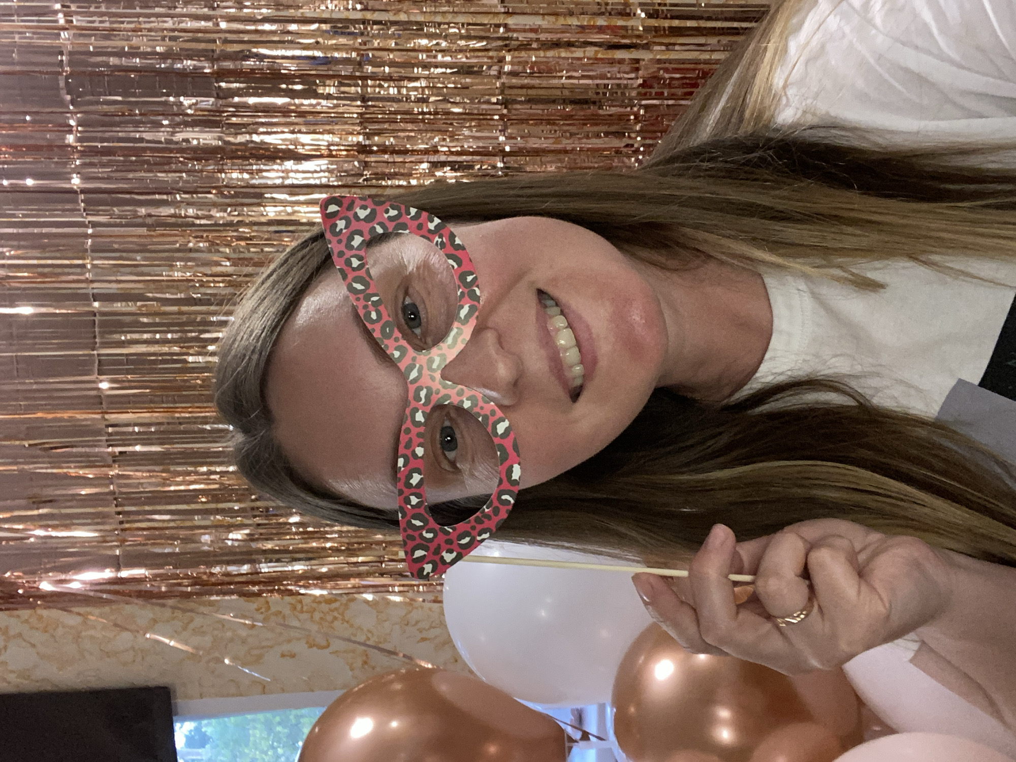 A fair-skinned woman with long, light brown hair is smiling and wearing novelty leopard-print glasses. She is holding the glasses' stick. The background consists of a metallic rose gold fringe curtain, and there are also rose gold and white balloons visible. The location is Chaikovsky, Russia.