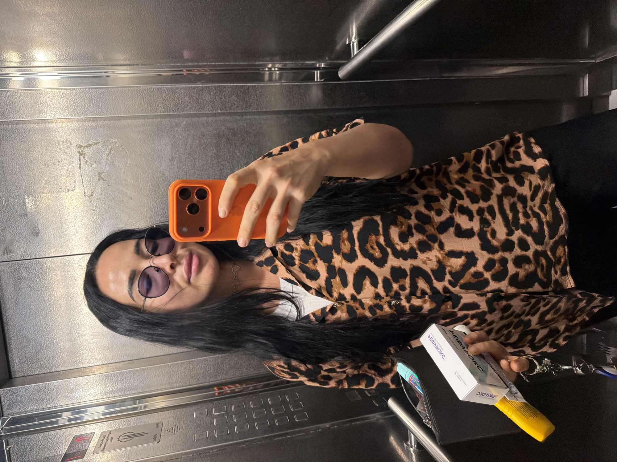A woman with long black hair and sunglasses is taking a selfie in an elevator in Saint Petersburg, Russia. She is wearing a leopard-print shirt over a white tank top and black pants. She is holding an orange smartphone in her right hand and a black clutch with a yellow object and a white box in her left hand. The elevator walls are made of brushed metal. The elevator doors are open on the right side, revealing a glimpse of the building's interior.