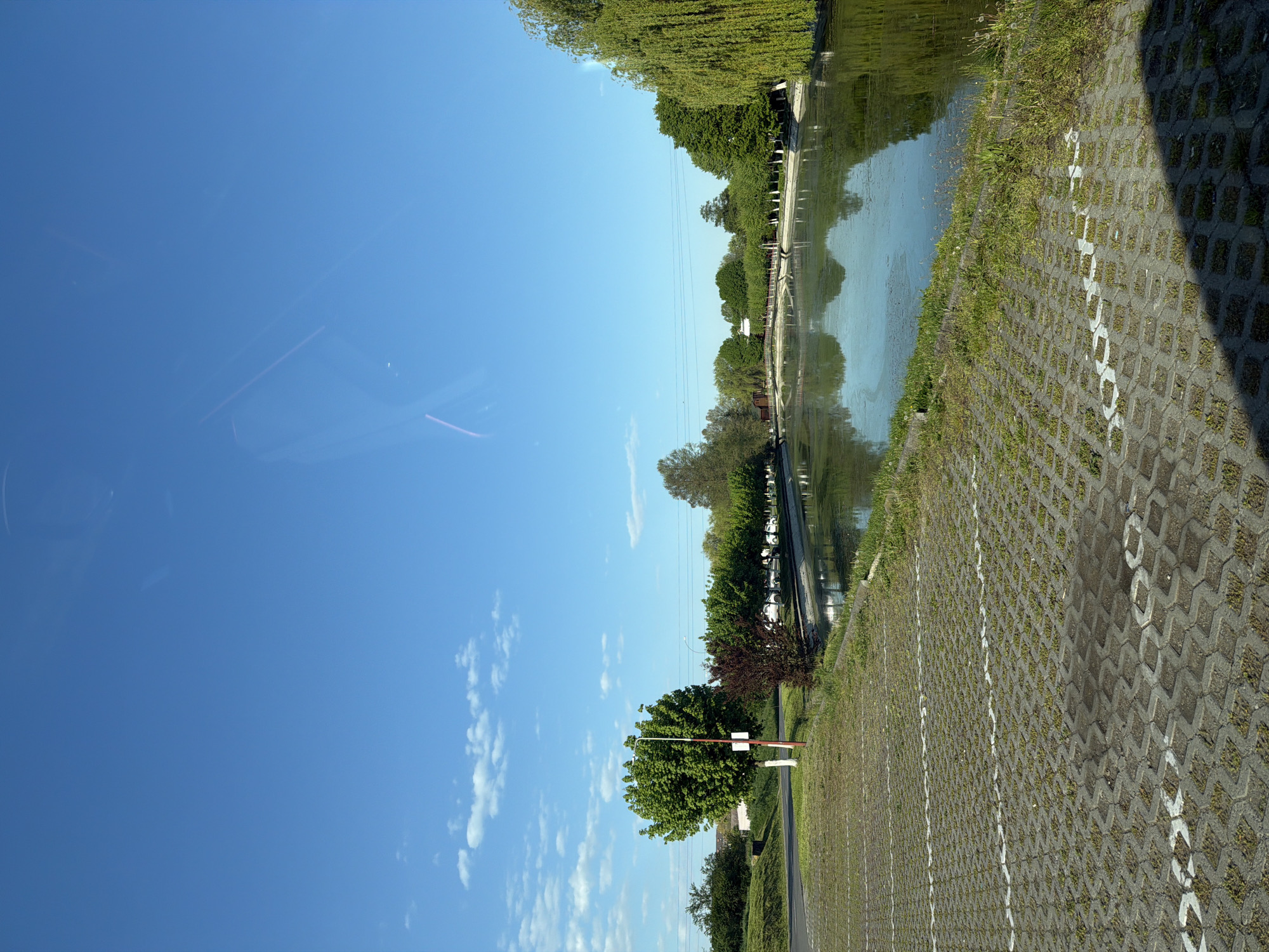 A low-angle landscape oriented view of a canal scene with a blue sky and clouds, taken in Târgu Mureș, Romania. The canal runs horizontally across the top right quadrant. Reflections of trees and the sky are visible in the water. The near side of the canal is bordered by grass and a grey block paved area, with white painted parking space markers. A red and white barrier and some trees are visible in the lower center of the image. The sky occupies a large area in the left side of the image. The view is from inside a vehicle, with some internal reflections visible.