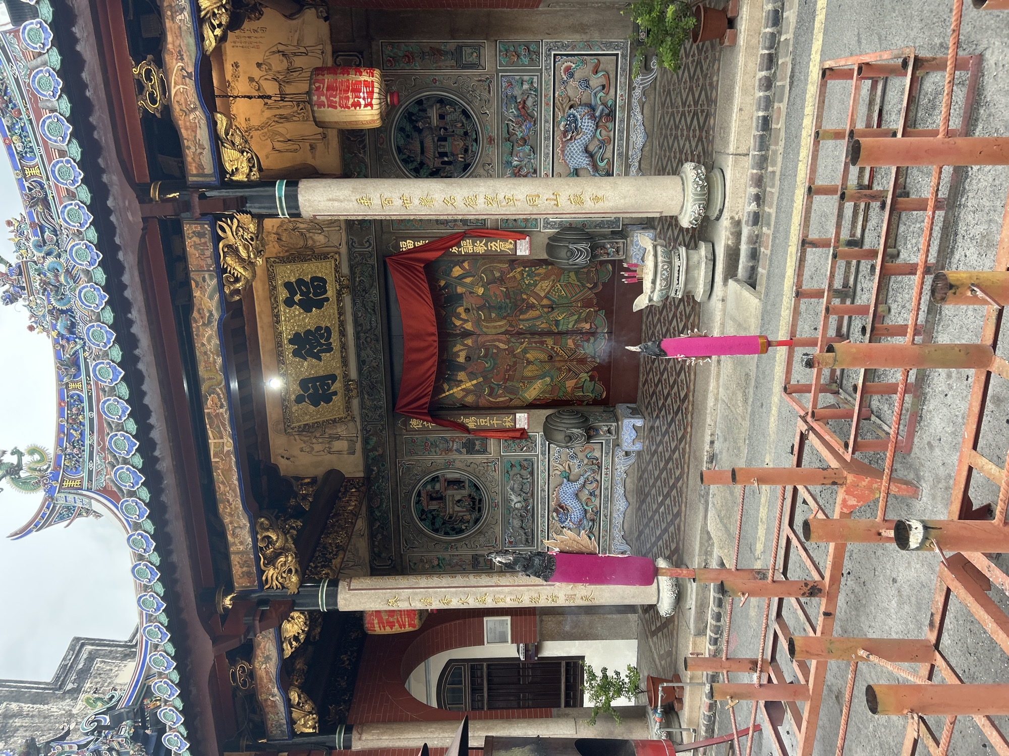 This is a medium shot, eye-level photograph of the entrance to a temple in George Town, Malaysia. The temple features ornate architecture with intricate carvings and vibrant colors. A red curtain hangs over a large, ornate wooden door. Two large pink incense sticks are burning in the foreground, placed in holders. In front of the temple entrance, there is a stone incense burner with several red incense sticks also burning. The temple is decorated with traditional Chinese motifs, including dragons and human figures. The ground in front of the temple is paved with patterned tiles. A metal barricade, painted orange, is situated in the foreground, partially obscuring the view of the temple's lower facade. The sky is overcast.
