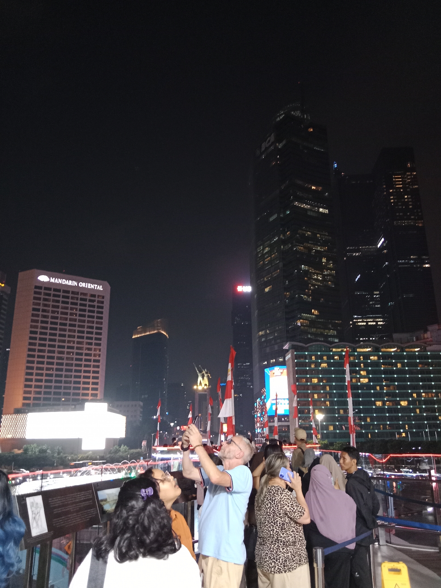 This is a nighttime outdoor photograph of a crowd of people gathered at an urban plaza in Central Jakarta, Indonesia, likely near the Monumen Nasional (Monas) area, given the presence of Indonesian flags. In the background, several tall skyscrapers are illuminated, including the Mandarin Oriental building with its name prominently displayed. To the right, a modern building with many illuminated windows suggests a hotel or office complex. The crowd is diverse, with individuals of various ages and ethnicities. Some people are taking photographs with their phones, while others are engaged in conversation. The atmosphere appears to be festive, possibly due to a national holiday or event, as evidenced by the numerous Indonesian flags displayed. The foreground shows a railing and some display panels, suggesting a public viewing area or an exhibition. The lighting is artificial, coming from the buildings, streetlights, and possibly event lighting.