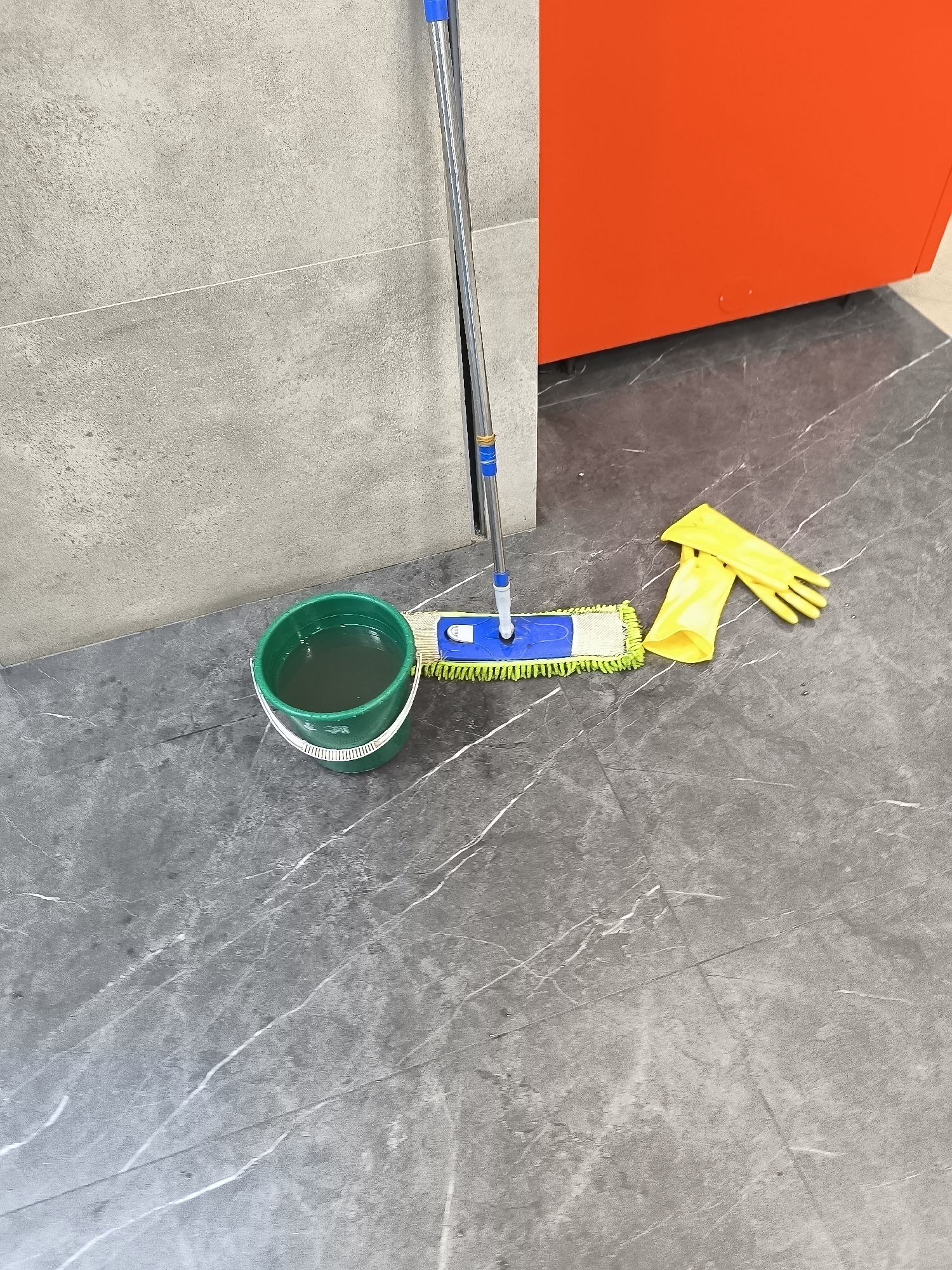 A green bucket filled with murky water sits on a gray marble-patterned floor next to a mop with a blue and yellow head and a silver handle. A pair of bright yellow rubber gloves lies to the right of the mop. In the background, a gray tiled wall and a bright orange cabinet are visible. The scene is devoid of human presence and appears to be a still-life arrangement of cleaning supplies.
