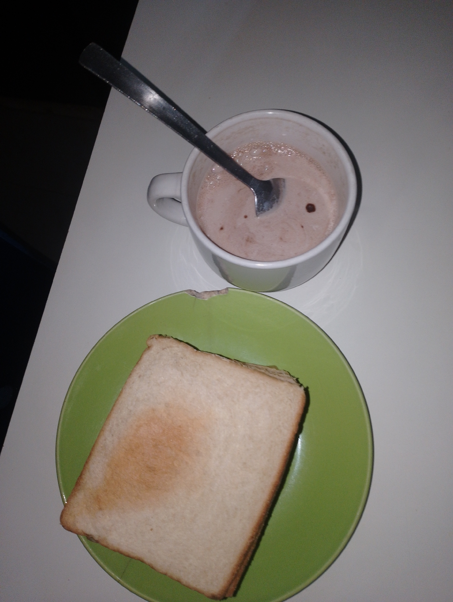 A cup of hot chocolate with a spoon in it sits on a white surface next to a green plate holding a slice of toasted bread. The scene appears to be indoors, likely in a home or café, on a white table. There is no indication of its location in Katon Rikkos, Nigeria. The lighting is dim, suggesting it might be evening or nighttime.