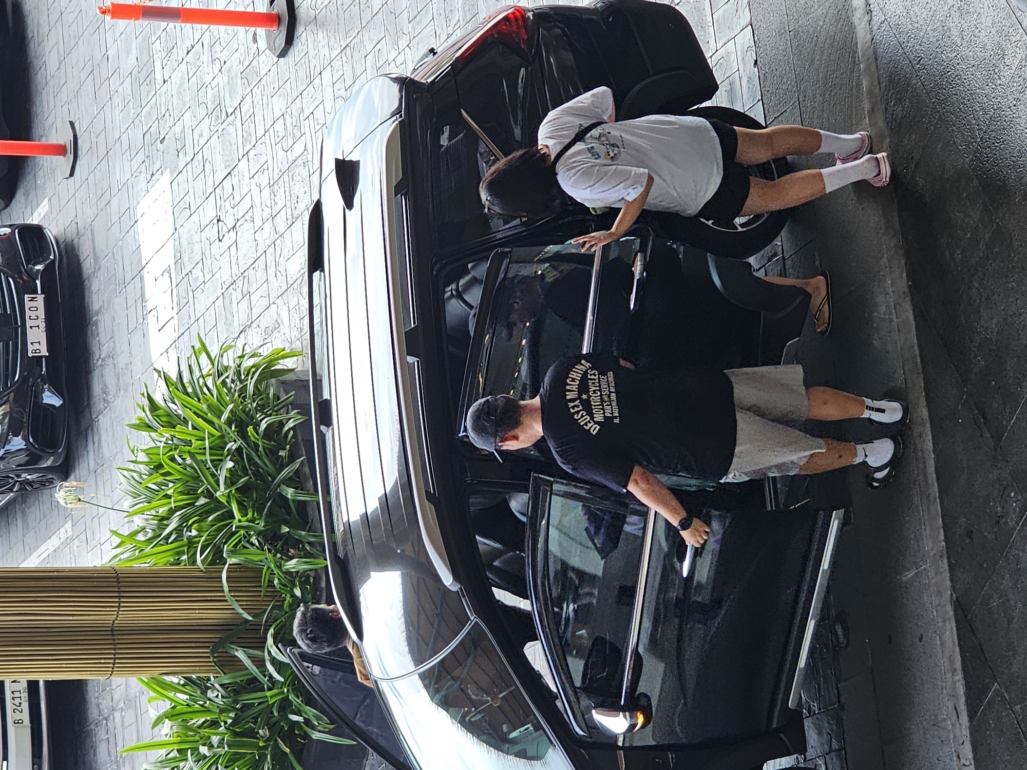 The image depicts a scene in Denpasar, Indonesia, where a black SUV is parked. Two individuals are near the opened rear passenger door. A person wearing a black t-shirt and grey shorts is standing by the rear driver's side door, with their hand on the handle, facing towards the rear of the vehicle. Another person, wearing a white t-shirt and black shorts, is facing the rear passenger door and appears to be interacting with someone inside or about to enter the vehicle. The ground is paved with interlocking stones, and there are green plants and a decorative bamboo column visible in the background, suggesting an urban or commercial setting. A silver car with a license plate starting with "B 2411" is parked in the upper left portion of the image. Red traffic cones are also present on the right side of the image.