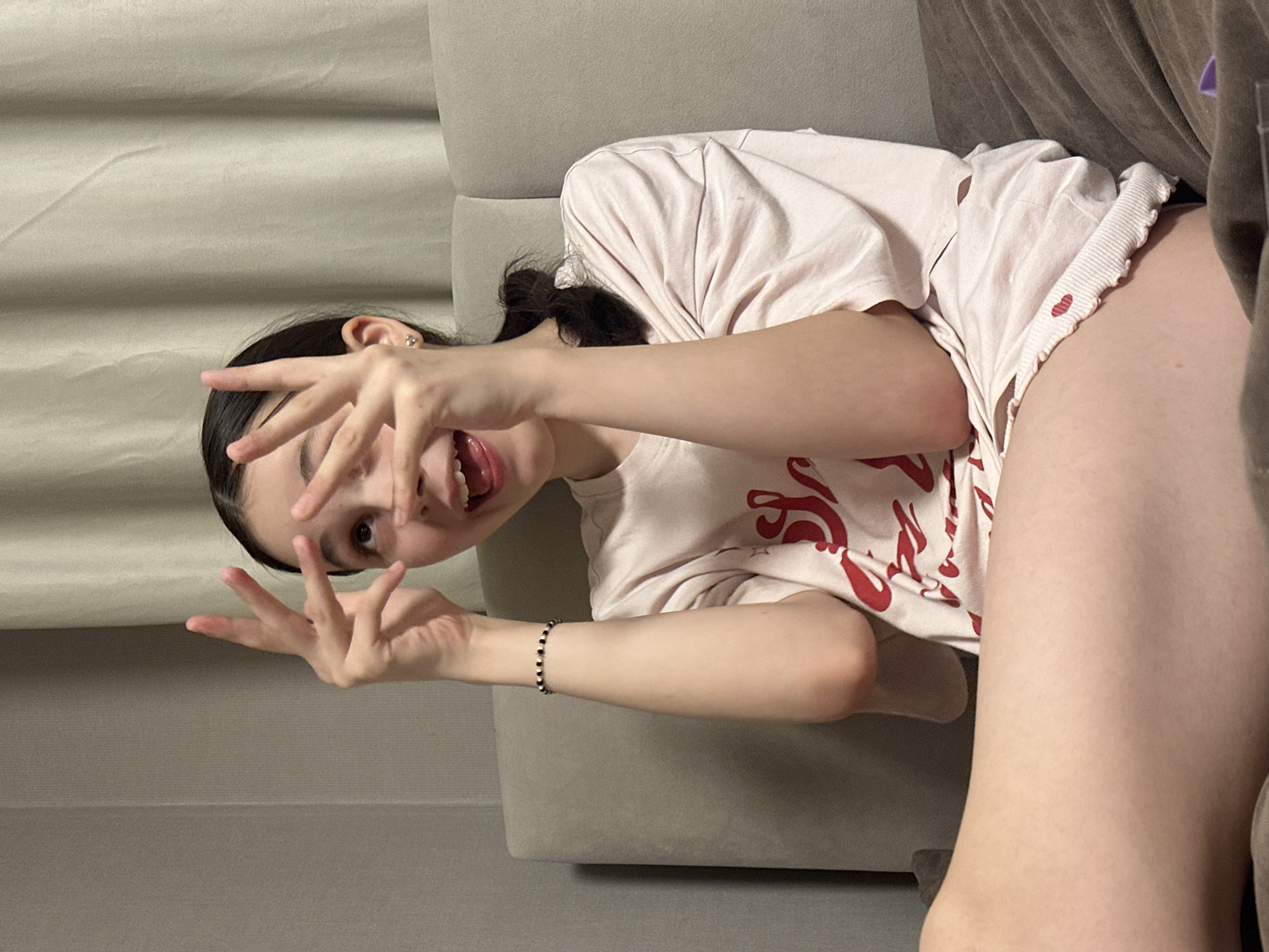 A young woman is reclining on a light gray couch, facing the camera. She is making a peace sign with both hands, one near her forehead and the other lower. Her hair is dark and pulled back. She wears a light pink t-shirt with a red pattern and matching shorts. The background includes a beige wall with horizontal lines. The location is unspecified, with no indication of Votkinsk, Russia.