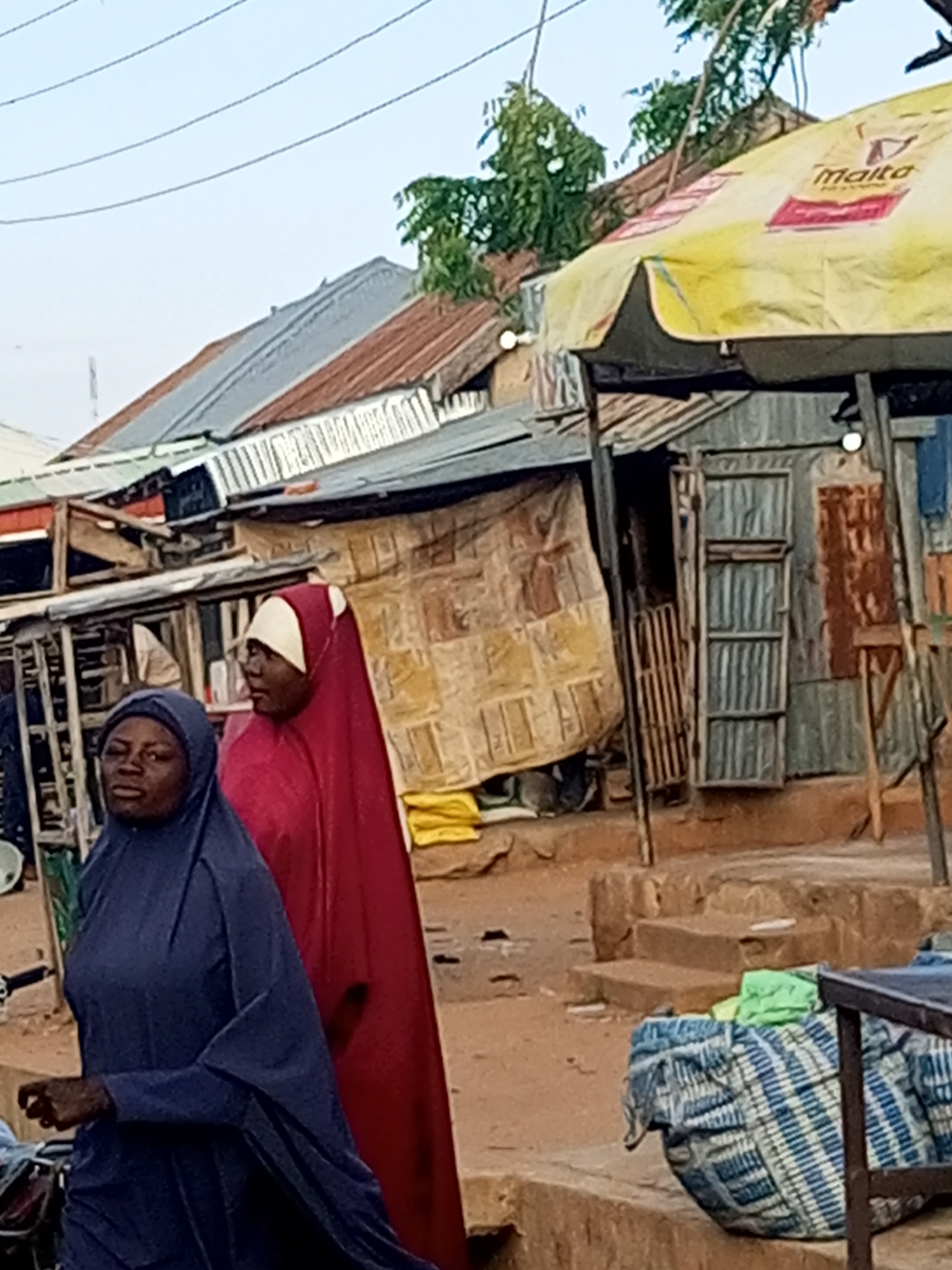 The image shows a street scene in Tempure, Nigeria. Two women are walking in the foreground, both wearing hijabs. The woman on the left is wearing a dark blue hijab, and the woman on the right is wearing a red hijab with a white cap. Behind them, there are several buildings made of corrugated metal and wood, with some covered by tarps. A yellow umbrella with the "Malta" logo is visible in the background. The ground is dirt and there are some steps leading up to one of the buildings. The overall impression is of a busy, informal market area.
