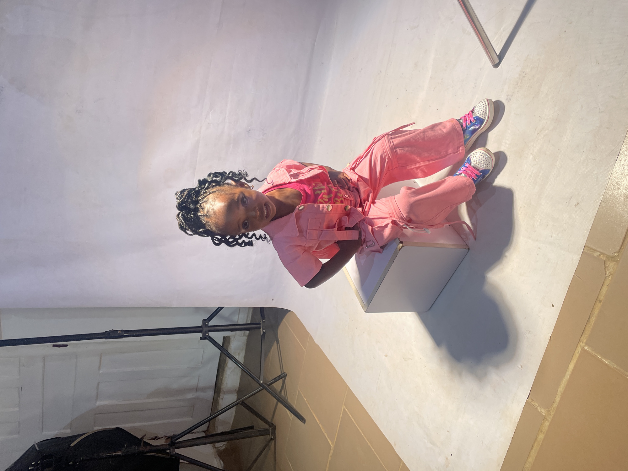A young girl with braided hair is sitting on a white cube in a studio setting. She is wearing a pink jumpsuit over a pink top and colorful sneakers. The studio has a white backdrop and floor, and photography equipment is visible in the background. The location is not explicitly evident in the image. Barakin Akawo, Nigeria cannot be confirmed as the location.