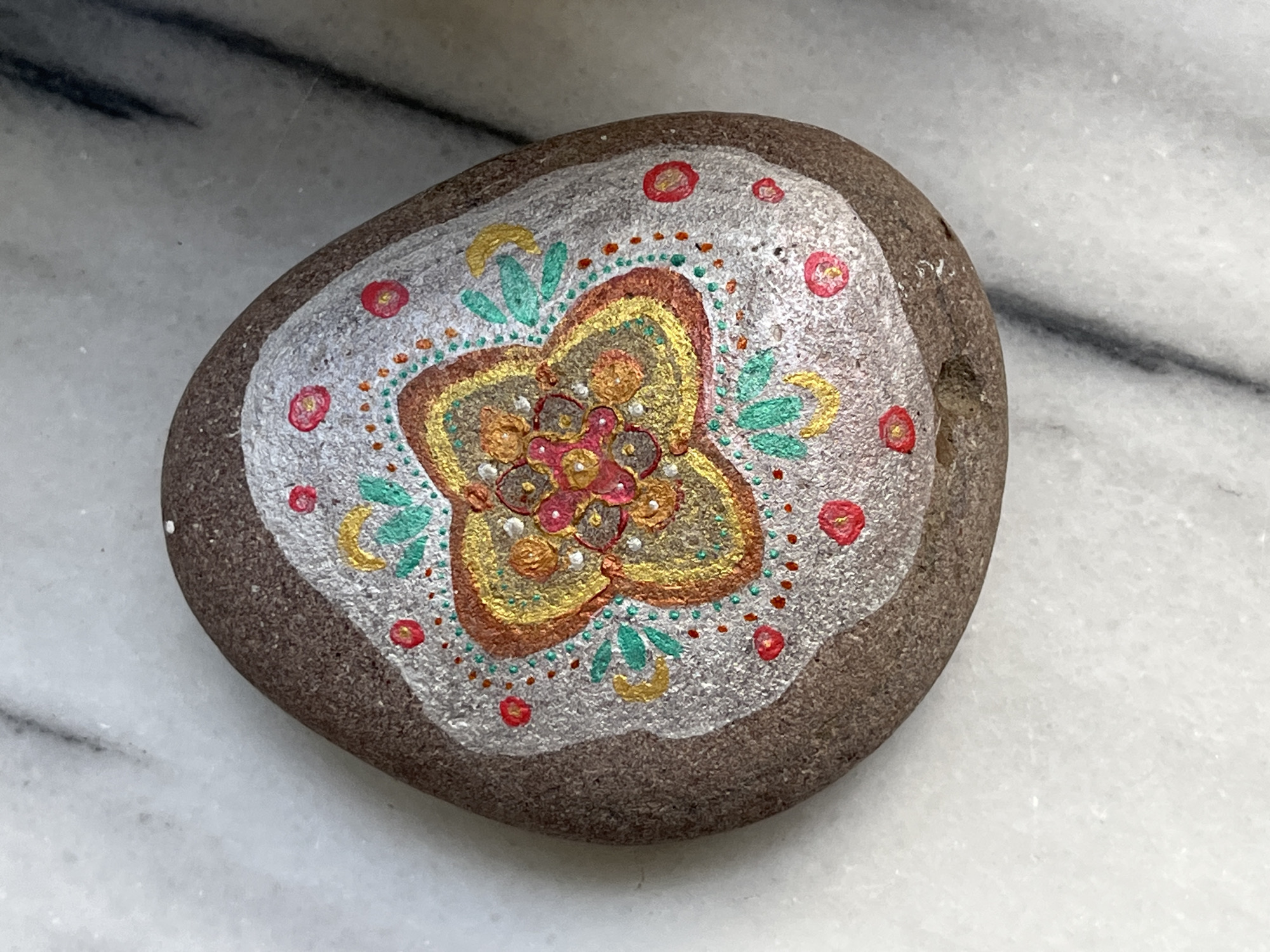 This media file shows a close-up, overhead view of a painted rock. The rock is oval-shaped and has a dark brown, speckled surface. It is resting on a light-colored, marbled surface.
The rock is decorated with a colorful, mandala-like design. The center of the design features a floral pattern with intricate details. The petals of the flower are painted in shades of gold, brown, and pink, with small white dots and red accents. Surrounding the central flower are various decorative elements. There are green leaf-like shapes, crescent moons painted in yellow, and small red circles with yellow centers. The entire design is set against a background of silver paint. The silver paint has a slightly textured appearance.
The lighting in the image is bright, suggesting it was taken during the day, possibly indoors with artificial light or near a window. There is no visible text in the image. The overall impression is of a handcrafted decorative object, likely created as a piece of art or a small gift. The marbled surface it rests on provides a neutral backdrop that highlights the detailed artwork on the rock.