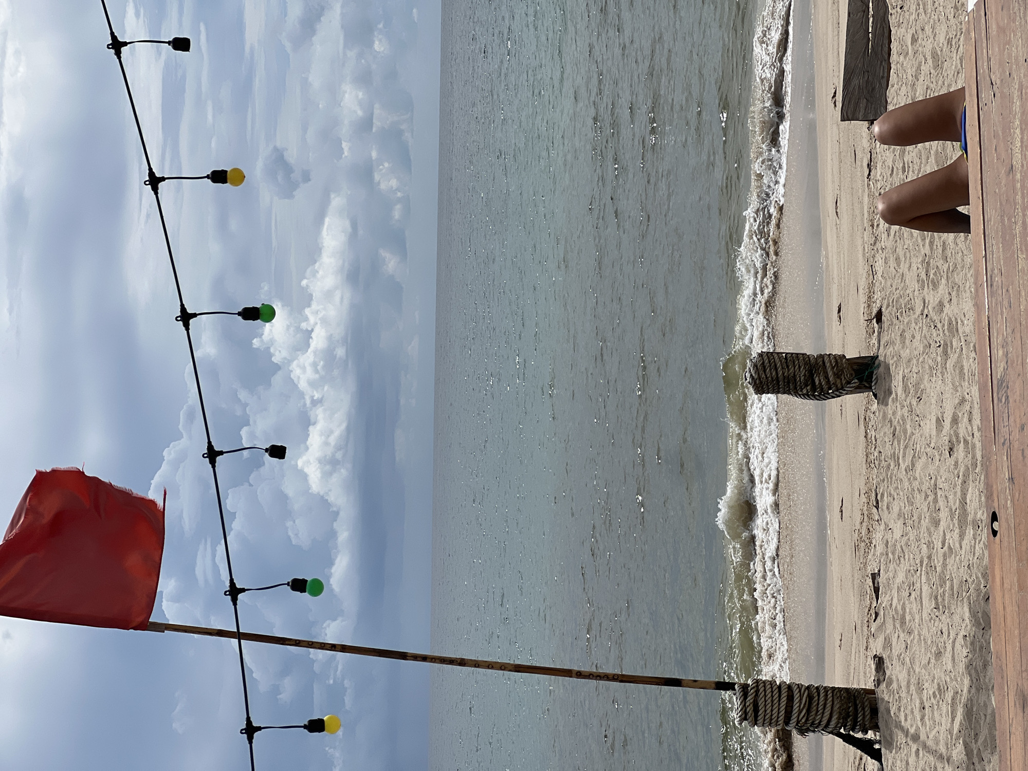 A red flag flies on a wooden pole at a beach. The flag is torn. Below the flag, a string of lights hangs across the scene, with some lights lit yellow and green. The ocean is visible in the background, with waves crashing on the shore. The beach is sandy, with two wooden posts wrapped in rope on either side of the foreground. In the foreground, the legs of a person are visible, lying on the sand. The person is wearing blue shorts. The scene is set against a cloudy sky.