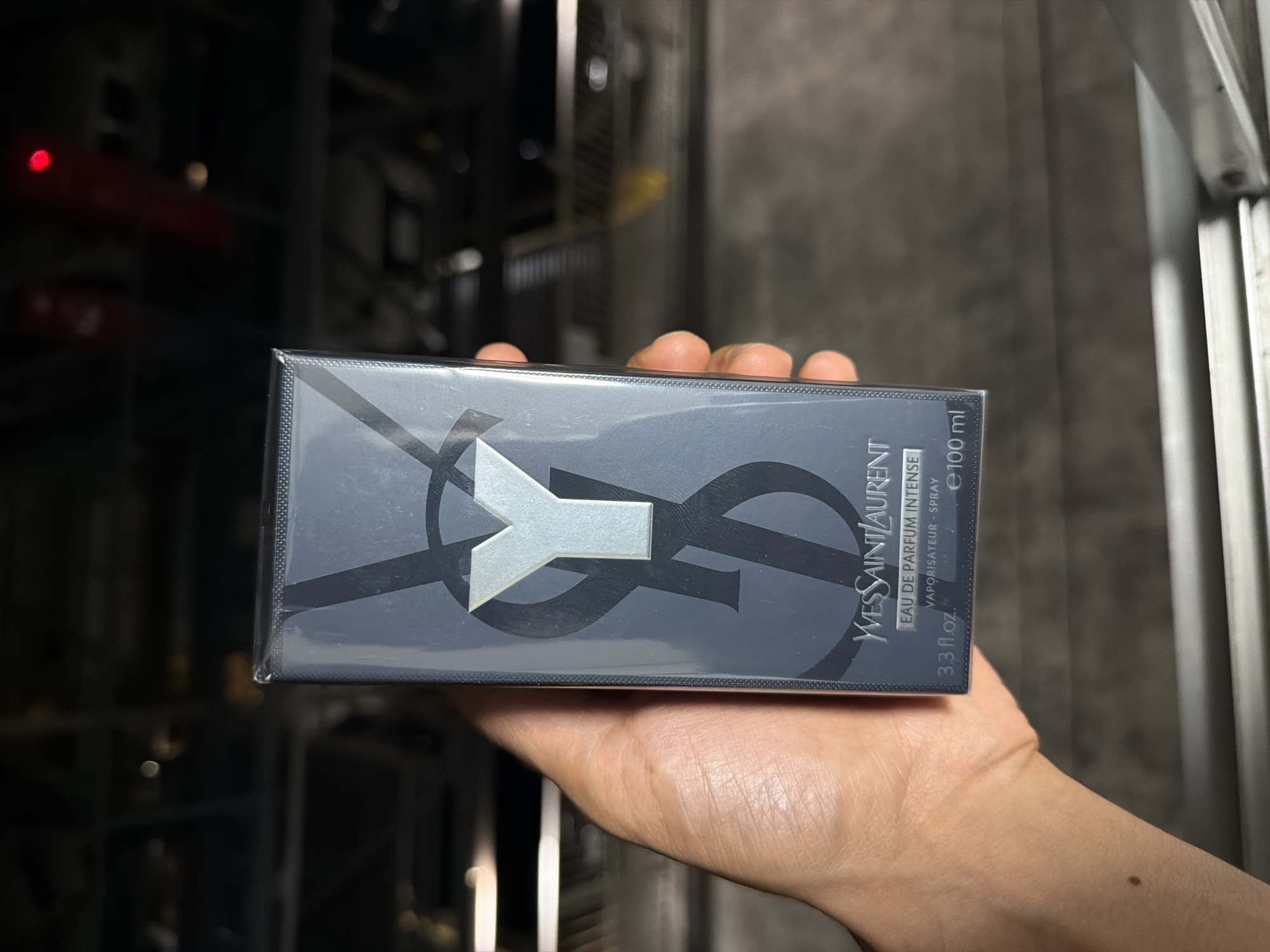 A hand is holding a boxed perfume. The box is dark blue with a large, stylized "Y" in silver on the front. The text on the box reads "YVES SAINT LAURENT EAU DE PARFUM INTENSE VAPORISATEUR - SPRAY 3.3 FL.OZ. e 100 ml." The background is dark and out of focus, suggesting an indoor, possibly dimly lit environment. There is a faint red light visible in the upper right portion of the background. The image is cropped close to the perfume box and hand, with the surrounding environment largely obscured. There is no indication of the specific location within Adachi, Japan.