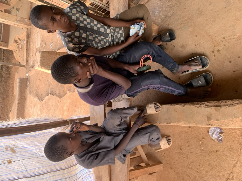 Three male juveniles are observed seated on a wooden bench within an outdoor or semi-sheltered area in Ube, Nigeria. The ground consists of uneven, reddish-brown earth.
The juvenile on the left wears a dark grey long-sleeved shirt and trousers with beige slide sandals, his right hand positioned near his mouth. The central juvenile wears a dark purple t-shirt and dark trousers with side stripes. He is barefoot, with dark slide sandals on the ground nearby, his right hand covering his mouth, and his left hand holding a green device with a red elastic band. The juvenile on the right wears a grey and dark checkered short-sleeved shirt and light brown trousers. He is barefoot, with dark slide sandals on the ground nearby, and holds a stack of Nigerian Naira banknotes in his left hand.
In the background, a light-colored striped fabric screen is visible on the left, and eroded earthen structures or steps are present towards the right. A small, translucent plastic bag containing unidentifiable contents rests on the ground in the foreground.