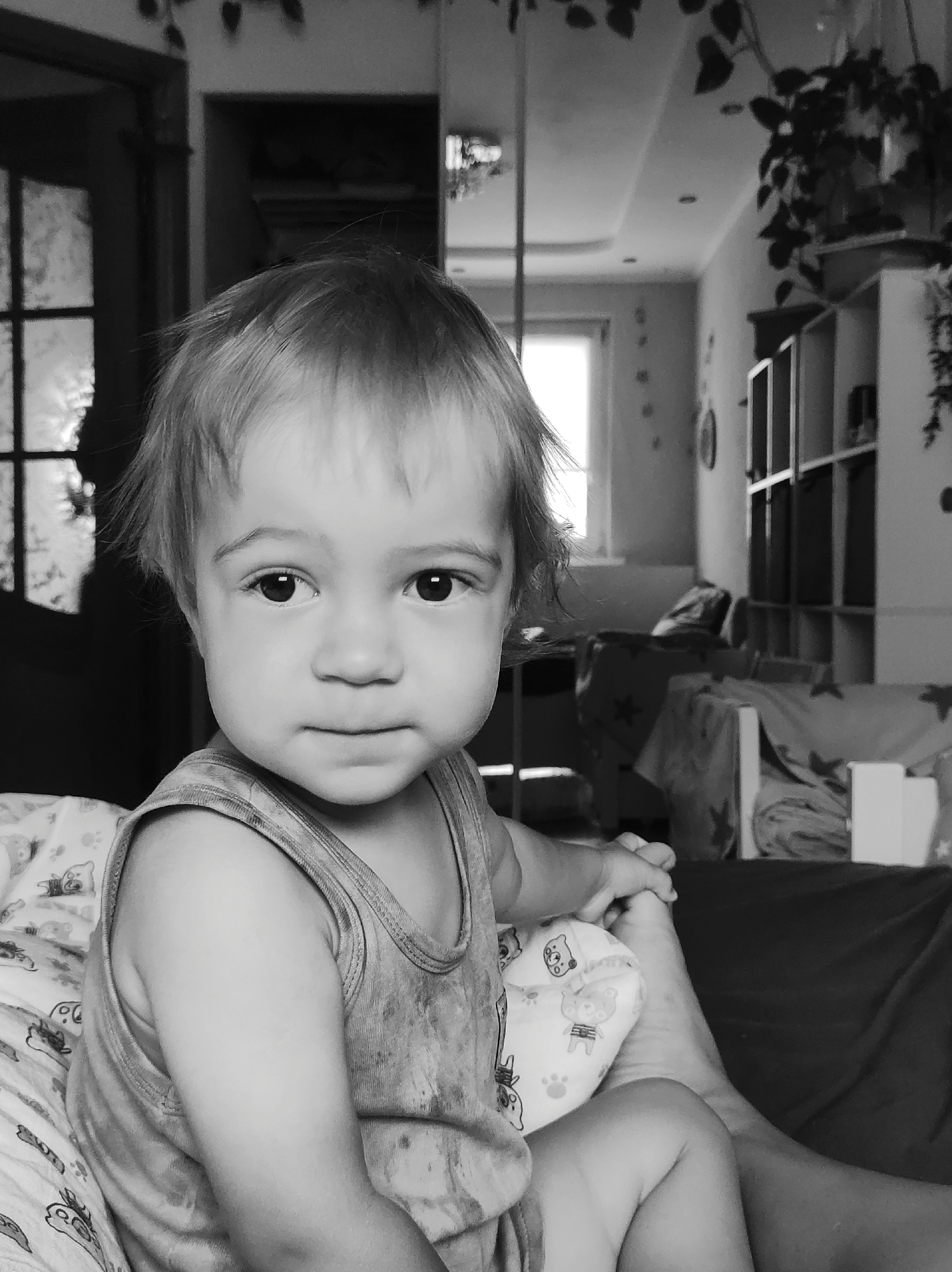 A black and white, medium close-up, waist-up, chest-up shot captures a fair-skinned toddler in the foreground, looking directly at the viewer. The toddler has short, light-colored hair, dark eyes, and is wearing a sleeveless tank top. The background appears to be an indoor domestic environment, possibly a bedroom or living area, with a window emitting natural light. Furniture and shelves are visible in the blurred background, along with decorative plants. The image was taken in Chaikovsky, Russia.