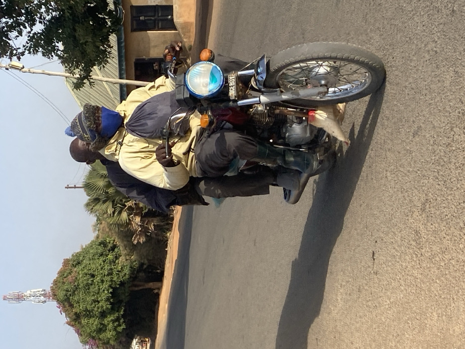 Two adult males are riding a red motorcycle on an asphalt road in Udu, Nigeria, under direct sunlight, casting long shadows to the left. The driver, wearing a light yellow jacket, dark trousers, dark green rubber boots, a blue face mask, and a knitted beanie with a blue pom-pom, grips the handlebars. A dark blue drawstring bag hangs from his neck. The passenger, wearing a dark jacket and trousers, sits behind the driver. The motorcycle has a round headlight, partially obscured by a light blue plastic covering, with "NICE BOY" inscribed below it, and orange turn signals. A white plastic bag is tied near the front wheel. In the background to the left, a large green tree with some red foliage stands, with a white cellular tower visible above it. To the right, additional trees and a light-colored building with a corrugated metal roof are present.