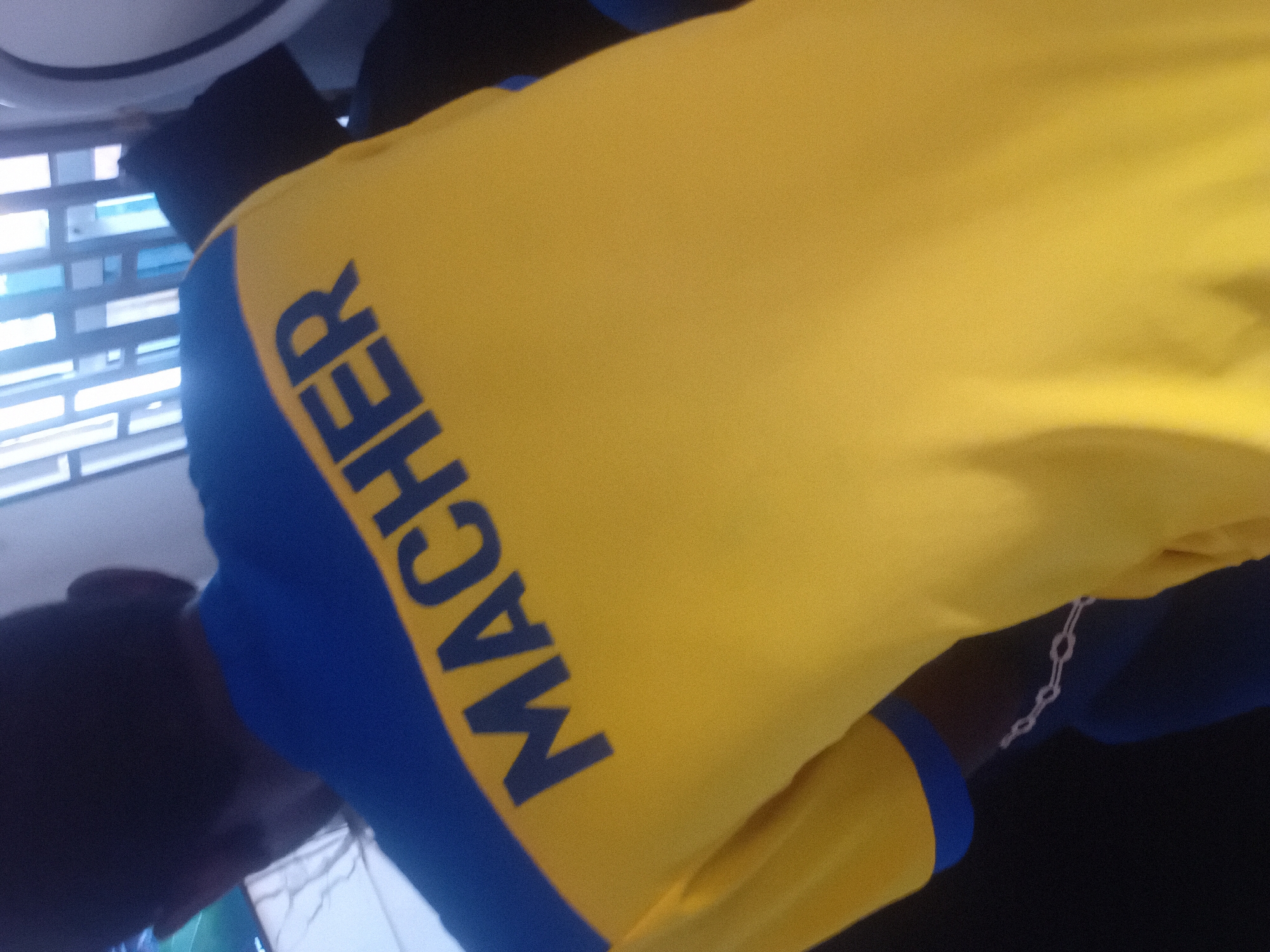 A person wearing a yellow shirt with blue trim is shown from behind. The word "MACHER" is printed in blue on the back of the shirt. The person's head and shoulders are visible. The background is blurry and appears to be an indoor environment with windows.