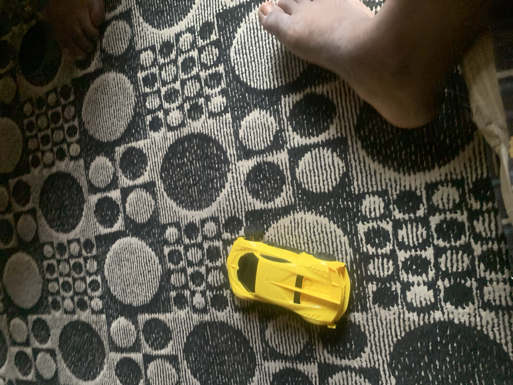 The image shows a yellow toy car positioned on a patterned rug with circles and squares. A person's feet are visible in the lower right and upper right corners of the image. The rug's pattern is predominantly black and white, with a textured appearance. The lighting suggests an indoor environment, possibly a home. The scene appears to be in Sabon Tasha, Nigeria, though no specific landmarks are visible to confirm this location. The focus is on the toy car and the rug.