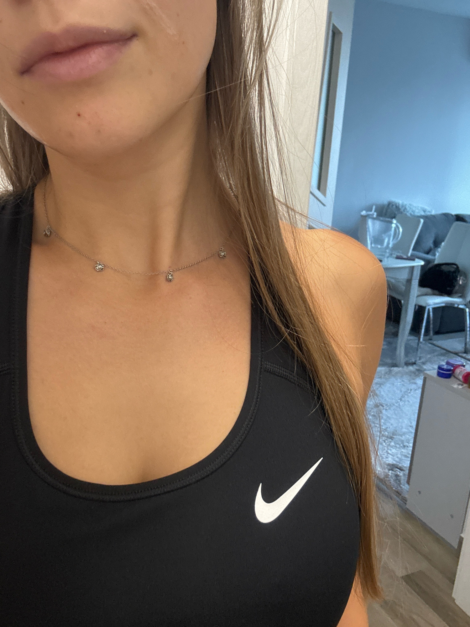 The image shows a close-up of a person from the chest up, wearing a black Nike sports bra and a delicate silver necklace with small charms. The person has long, light brown hair that falls over their shoulder. The background is blurred, but appears to be an indoor setting with furniture and a wall. No specific location in Sofia, Bulgaria is identifiable from the image.