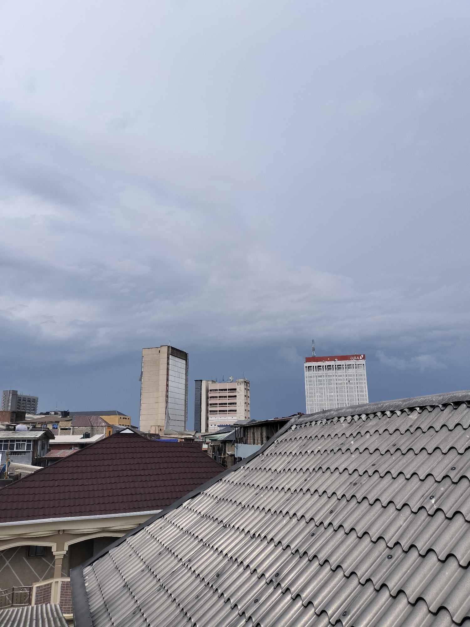The image shows a view over rooftops in Lagos, Nigeria. The foreground is dominated by corrugated roofs, one gray and one brown. Behind the rooftops, there are several buildings of varying heights, including at least three high-rise structures. The building on the right appears to have the letters "UBA" on top. The sky is overcast with dark, ominous-looking clouds.