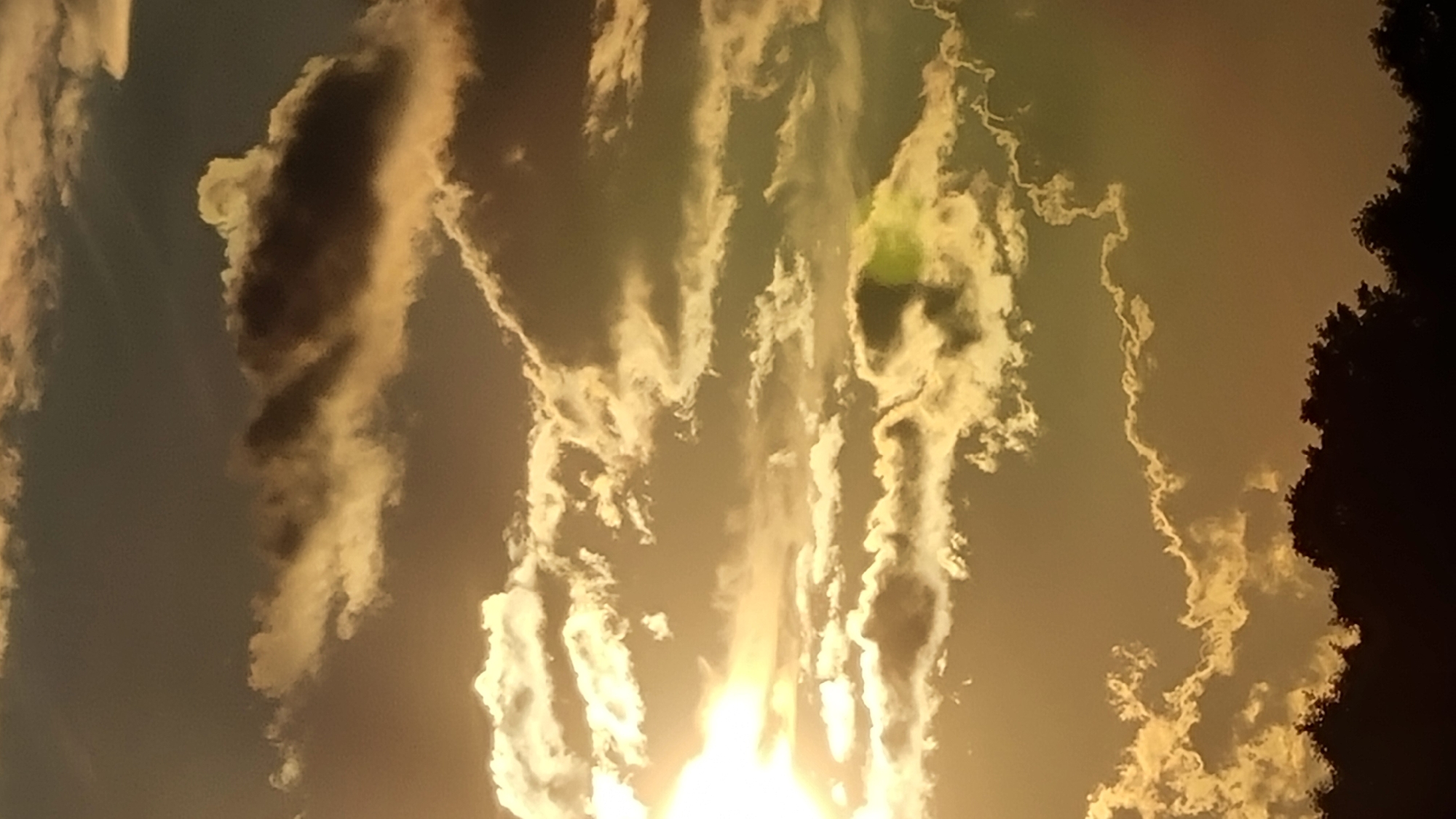 The image captures a dramatic sky filled with clouds, possibly during sunset or sunrise, casting a warm, golden light across the scene. The clouds exhibit a range of textures and densities, from wispy formations to dense, shadowed masses. In the lower portion of the image, the dark silhouettes of trees form a distinct horizon line, suggesting an outdoor setting. The overall mood is serene and expansive, dominated by the natural beauty of the sky. No people or specific urban features are visible. The location is stated to be Vysokoe, Russia.