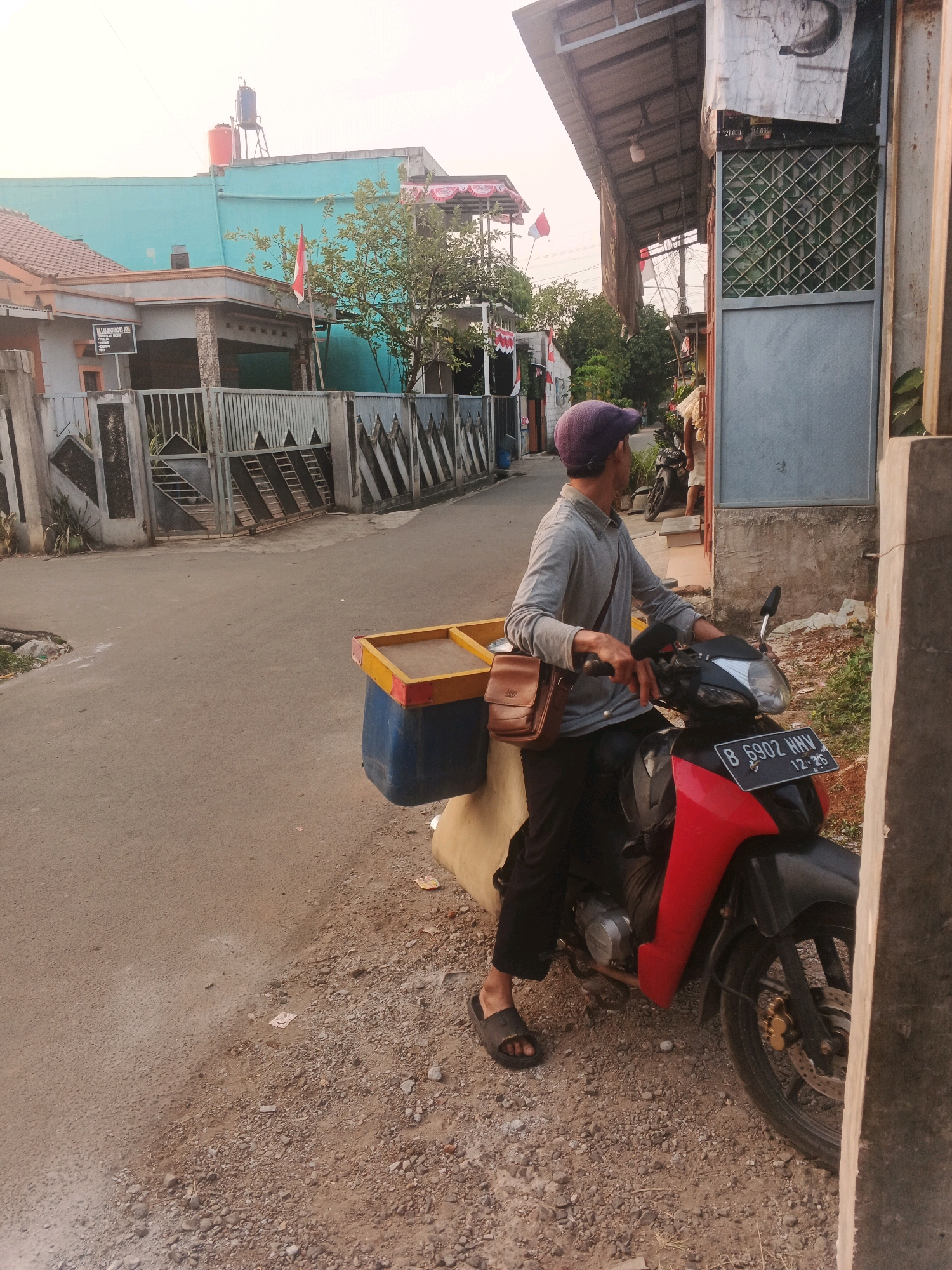 A man wearing a purple cap, grey long-sleeved shirt, black pants, and black sandals is sitting on a red and black motorcycle. He has a brown satchel slung across his chest. The motorcycle has a license plate reading "B 6902 HNW". Attached to the back of the motorcycle is a blue and yellow container. The scene appears to be a street in Kadu, Indonesia. The background shows residential buildings, a large metal gate, and trees. The sky is bright and appears to be late afternoon or early morning. The road is paved and there is gravel on the side of the road.