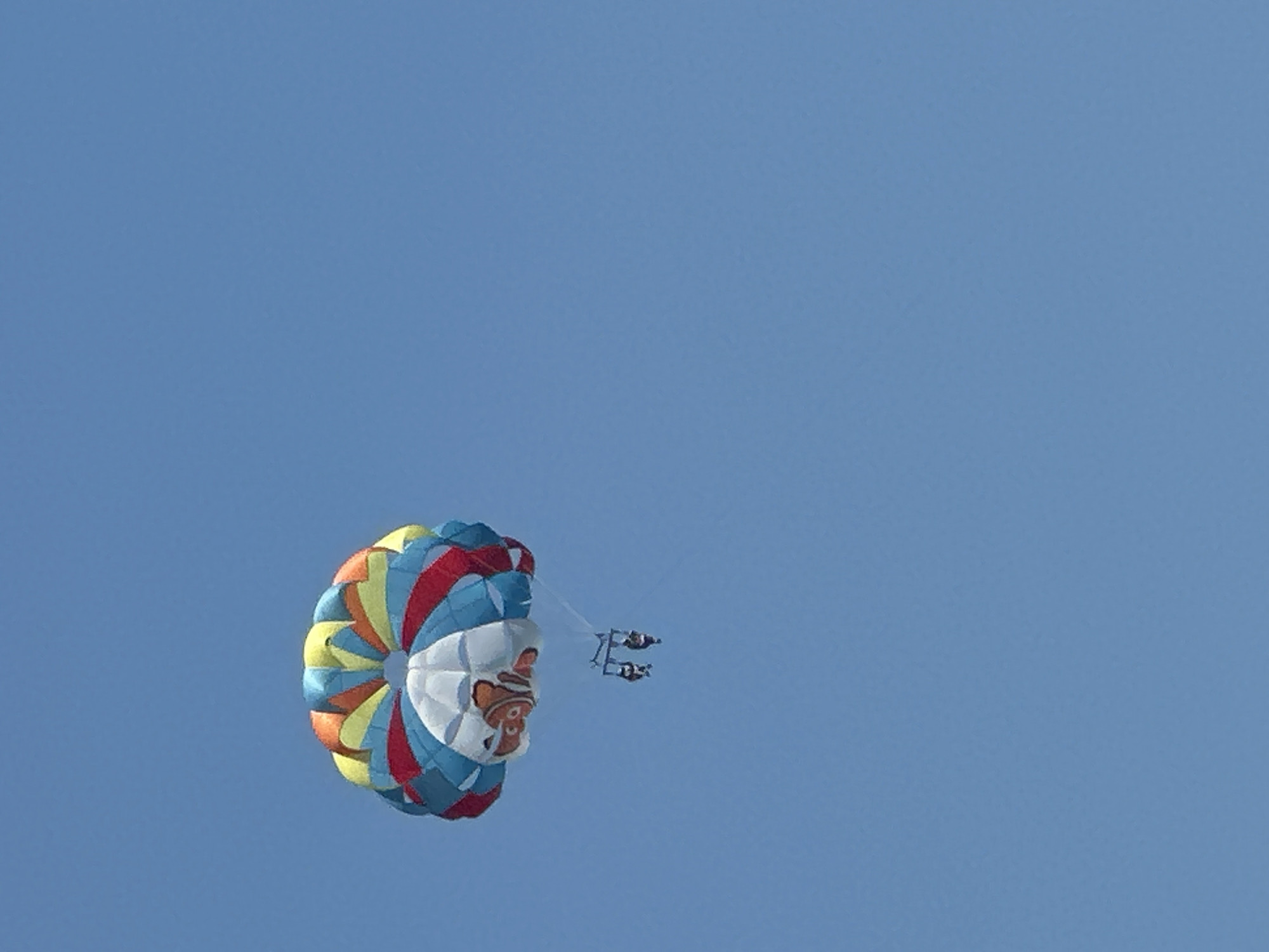 The image depicts a paraglider with a colorful parachute in mid-air against a clear blue sky. The parachute is predominantly white with red, blue, yellow, and orange sections forming a circular pattern. A cartoonish image of a clownfish, resembling the character Nemo, is visible on the white section of the parachute. Two people are suspended beneath the parachute, attached by harnesses and ropes, appearing to be enjoying the activity. The scene is likely a recreational activity taking place outdoors on a bright, sunny day, however, there is no information to determine the exact location in Boreti, Montenegro.