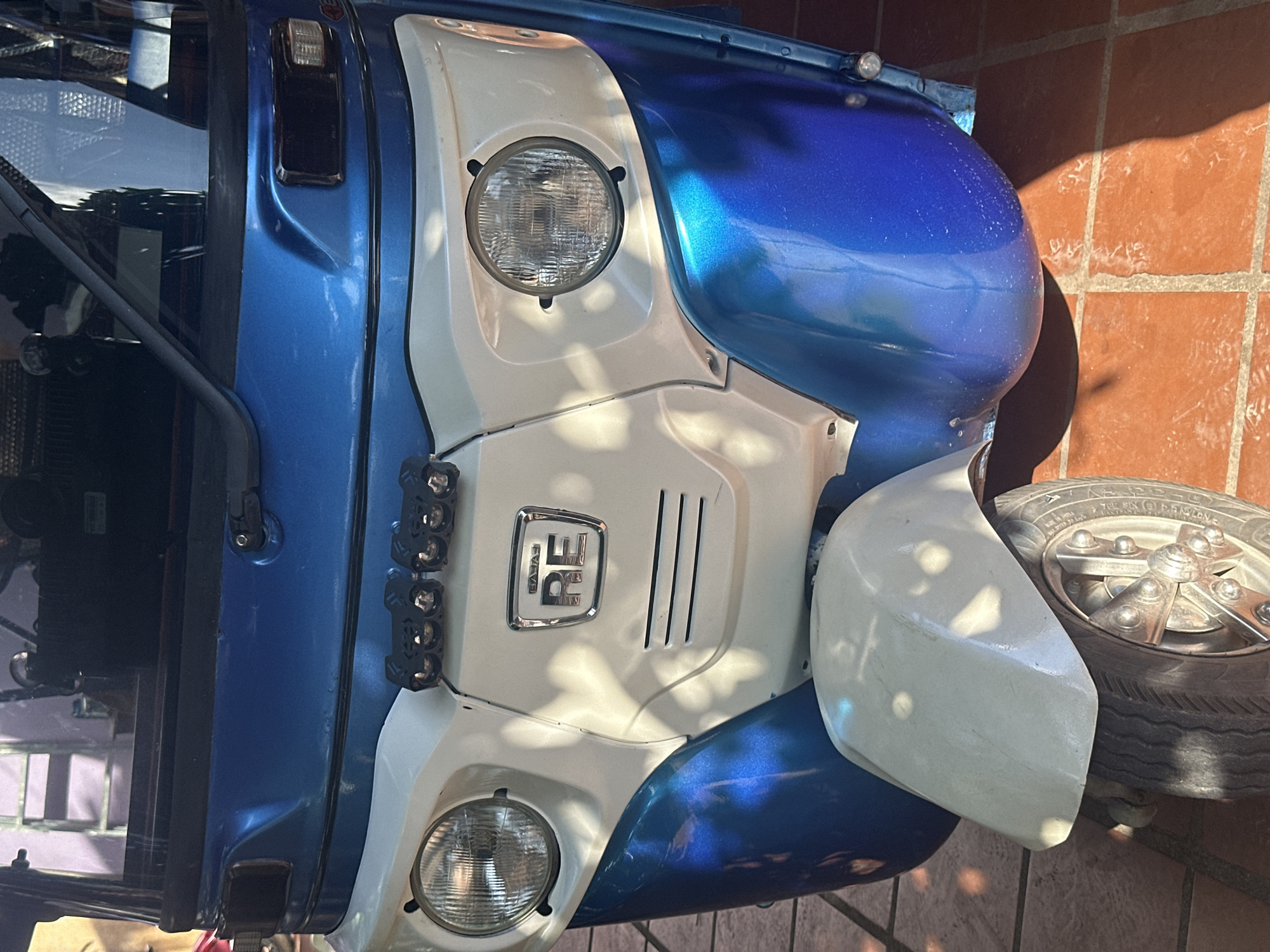 A close-up, eye-level shot shows the front of a blue and white three-wheeled vehicle, possibly an auto-rickshaw, parked on a tiled outdoor surface. The vehicle's front features a prominent white panel with a chrome emblem displaying "RE" in the center. Two round headlights are positioned on either side of this panel, and above them, a bar of four small, black auxiliary lights is mounted. The blue bodywork of the vehicle curves around the front, with a lighter blue metallic sheen visible. A section of the white front fender is visible above the front tire, which has a silver-colored hub. The ground is covered with reddish-brown tiles, some of which show a light sheen from sunlight and shadows. The background is out of focus but suggests an indoor or covered outdoor area with some structural elements. The image was taken in Kampot, Cambodia.