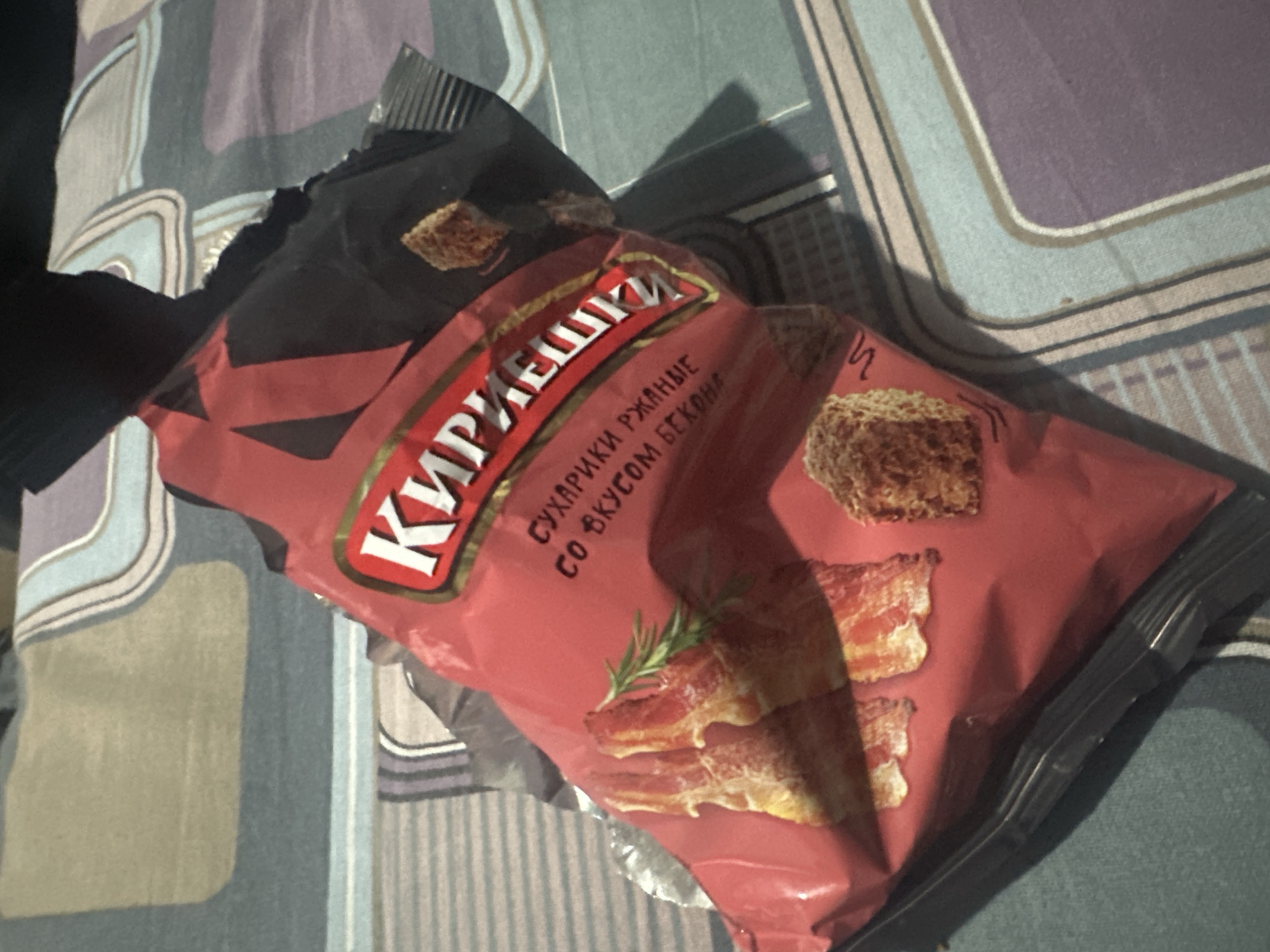 The image shows an opened bag of Kirieshki rye croutons with bacon flavor. The bag is red with black accents and features images of bacon and croutons. The text on the bag is in Russian. The bag is resting on a bedspread with a patterned design. The scene does not indicate the location within Gorno-Altaysk, Russia.