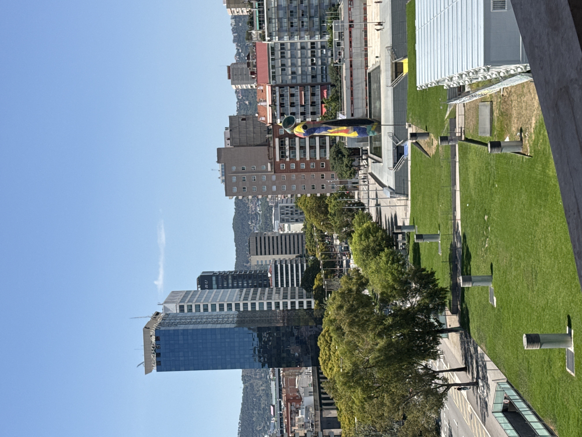This is a medium-shot, eye-level, outdoor, daytime photograph of a cityscape in Barcelona, Spain. In the background, several modern skyscrapers dominate the skyline, including a prominent blue glass skyscraper with a white structure on top, and a light-colored building with a grid facade. Behind these buildings, a hazy, mountainous landscape is visible. In the midground, a colorful, abstract sculpture stands tall, resembling a stylized human figure with a conical head and brightly colored mosaic patterns. In front of the sculpture, a large, rectangular pool of water reflects the blue sky. Surrounding the pool are tiered stone steps and walkways. To the left of the sculpture, a tree-lined street with parked cars is visible. The foreground features a lush green lawn with several cylindrical ventilation shafts protruding from the ground. A modern building with a white, corrugated metal roof is situated on the right side of the frame, partially obscuring the view of the lawn. A person is riding a bicycle on a paved path to the left of the lawn. The sky is clear and blue with a faint white cloud. The overall scene depicts an urban environment with a mix of modern architecture and public art.
