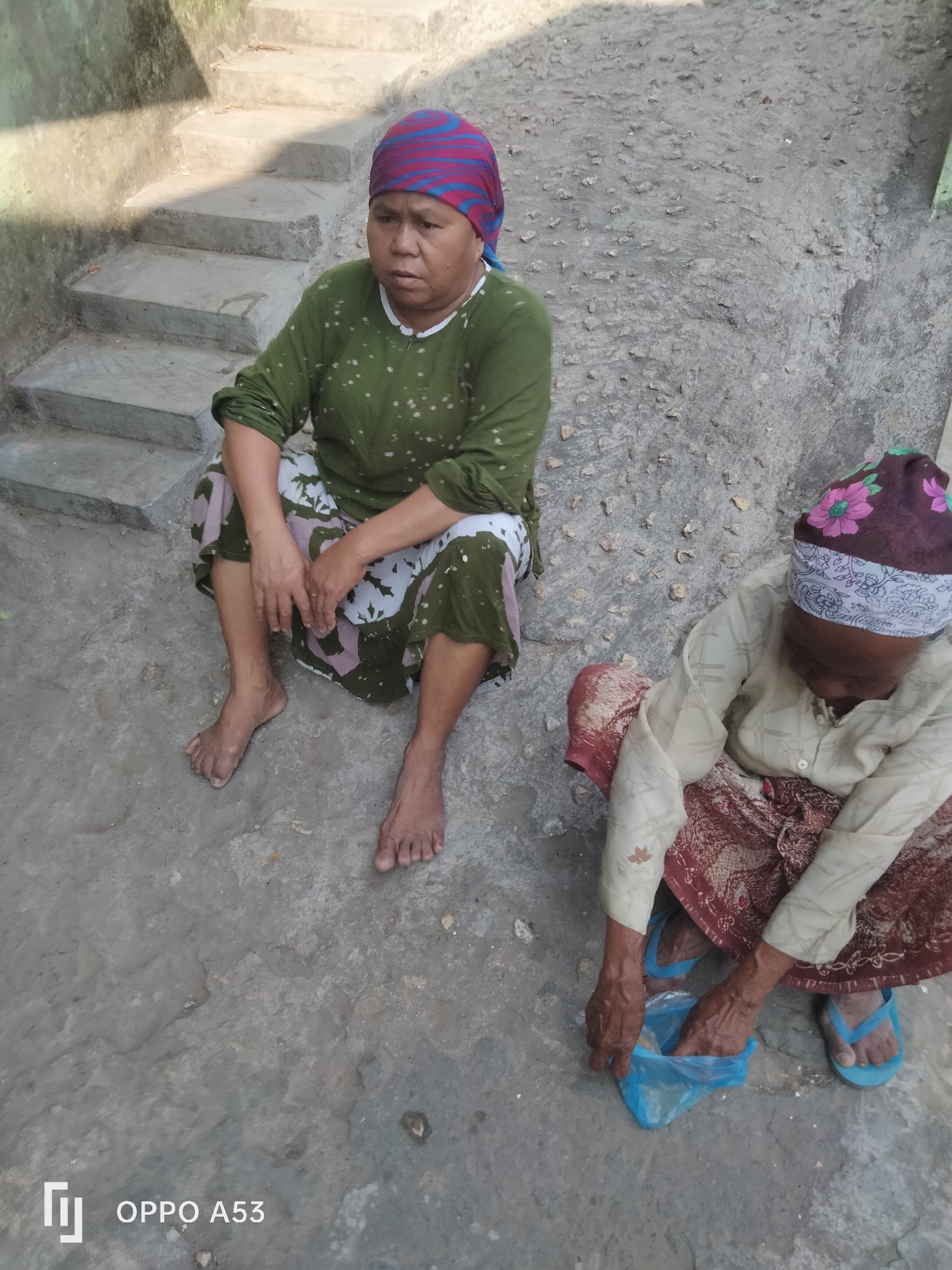 Two elderly Indonesian women are sitting on a concrete ground next to some stone steps. The woman on the left is wearing a green long-sleeved shirt with a patterned skirt and a headscarf with blue and red stripes. She is sitting with her legs extended and her arms resting on her lap. The woman on the right is crouching down and appears to be putting something into a blue plastic bag. She is wearing a light-colored long-sleeved shirt and a patterned skirt, and her head is covered by a headscarf with floral patterns. She is wearing blue flip-flops. The environment appears to be outdoors, possibly in a residential area of Badung, Indonesia, given the architecture and the presence of stairs and concrete pathways. The lighting suggests it is daytime. The image is sharp and clear, allowing for detailed observation of the subjects and their surroundings.