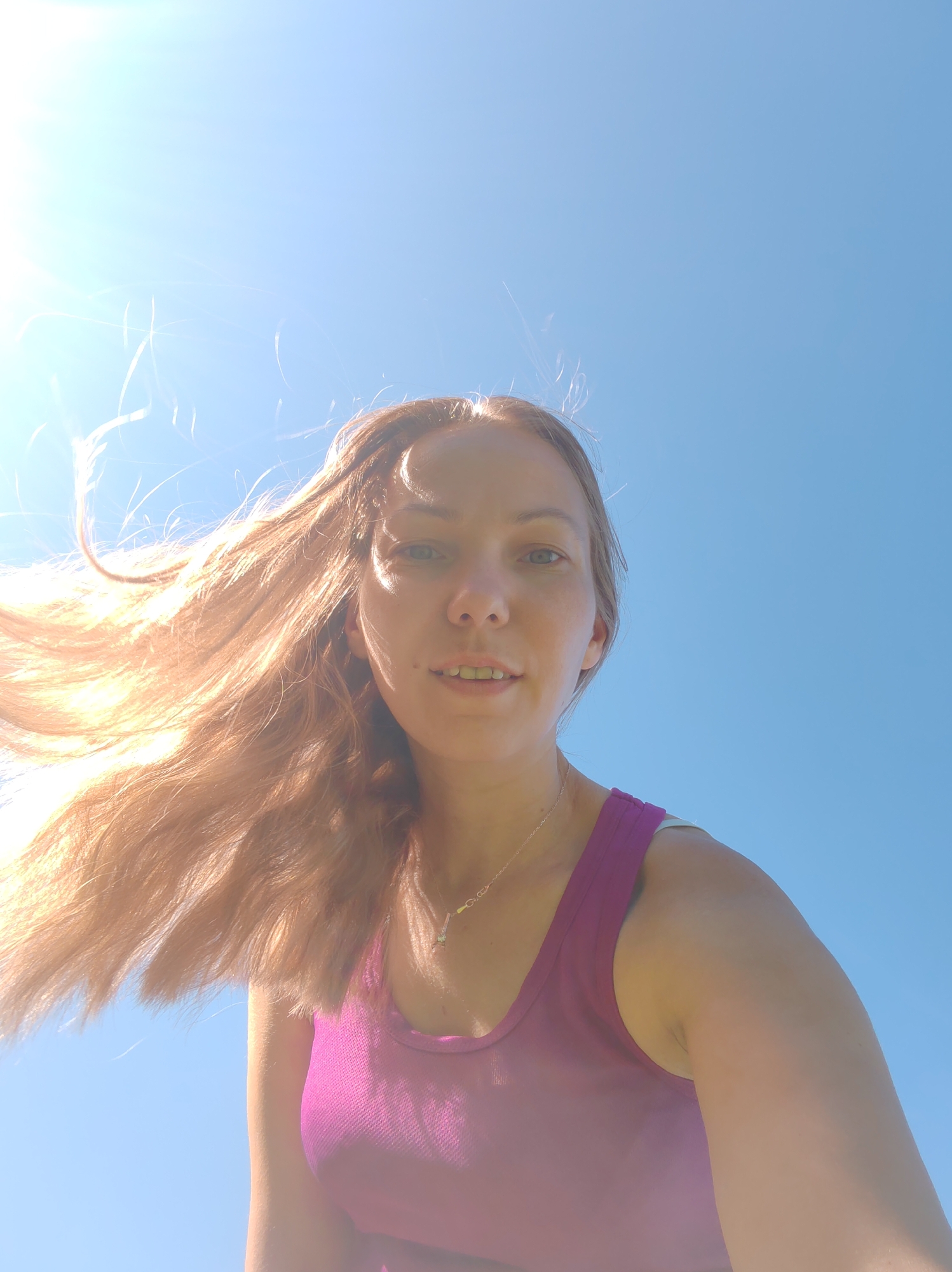 A low-angle, medium shot captures a young Caucasian woman from the chest up, looking directly at the camera. She has long, light brown hair that is being blown to her left by the wind, creating a dynamic effect against a bright, clear blue sky. The sun is visible in the upper left corner, causing lens flare and casting a bright light on her hair and face. She is wearing a bright magenta tank top, with a hint of a white strap from an undergarment visible. A thin, delicate necklace with a small pendant is worn around her neck. Her face is illuminated, with subtle makeup. The background is entirely the sky, indicating an outdoor setting, potentially a park or open area within the city of Chuiklem, Russia, given the clear, expansive sky.