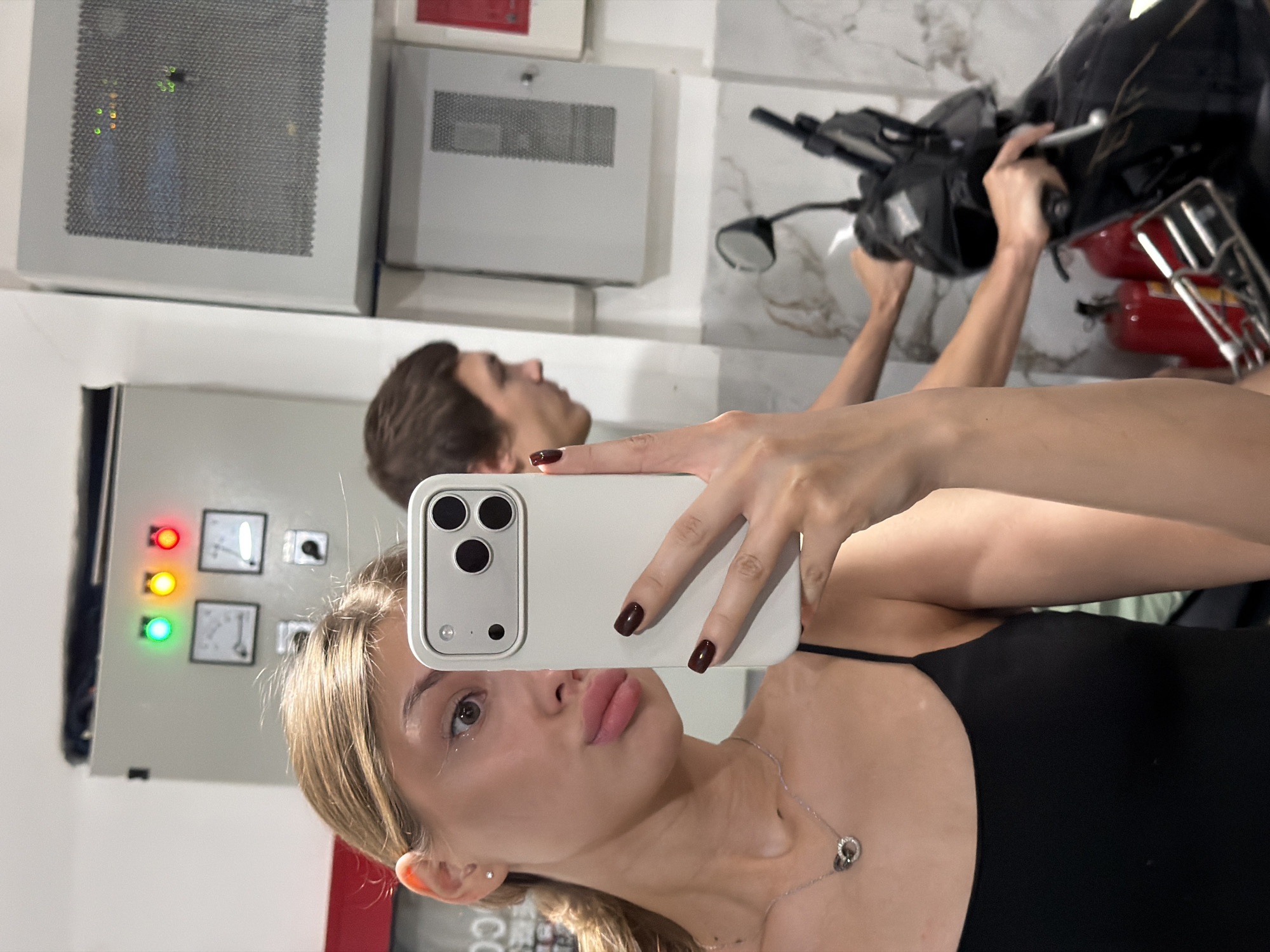 In a vertical, slightly tilted close-up shot, a young woman takes a selfie in a room that appears to be a garage or workshop, indicated by the presence of a motorcycle and industrial electrical panels. She is holding a white smartphone in her right hand, with her thumb resting on the screen. Her left arm is visible, bent at the elbow, and her left hand is also holding onto the motorcycle's handlebars, suggesting she is either seated on or near the motorcycle.
The woman has blonde hair and is wearing a black tank top and a silver necklace with two intertwined rings. Her nails are painted a dark, glossy color. Her expression is somewhat pouty, with her lips slightly parted. The background shows a large electrical panel with three colored indicator lights (red, yellow, and green), two analog gauges, and various switches. To the left, a metal ventilation grille is mounted on the wall, with some subtle reflections visible on its surface. The overall lighting suggests it could be daytime, possibly indoors, as there are no strong indications of natural light. The reflection in the electrical panel shows a young man in the background, also looking towards the camera. The presence of industrial equipment and a motorcycle points towards a functional space rather than a typical domestic setting. The scene is set in Đà Nẵng, Vietnam.