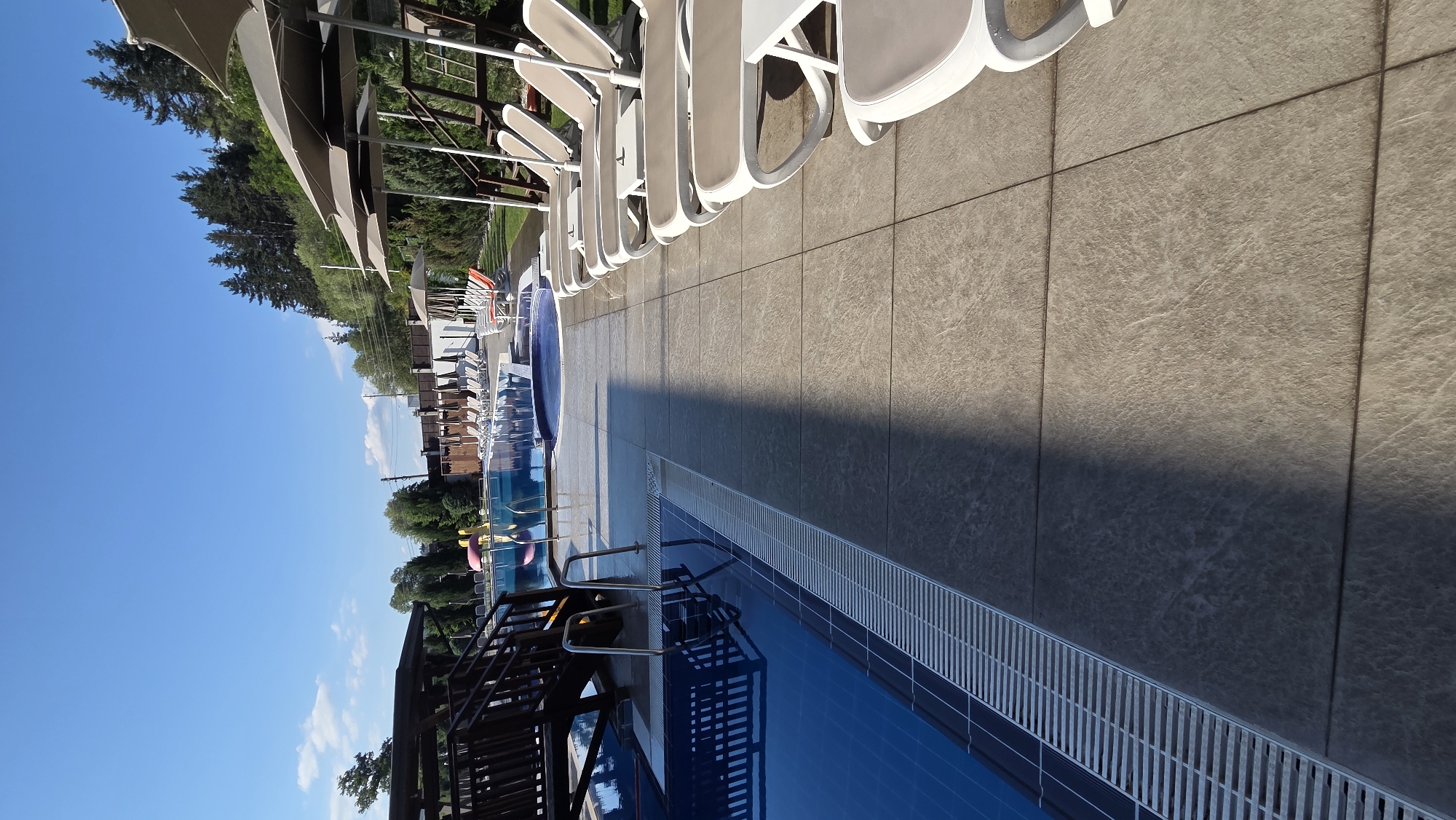 The image shows an outdoor swimming pool area with lounge chairs and umbrellas under a clear blue sky. The pool is blue and appears to be empty. There is a wooden structure with stairs next to the pool. In the background, there are trees and a fence. The area is paved with large, light-colored tiles. The image is a photograph of a resort in the city of Bania, Bulgaria.