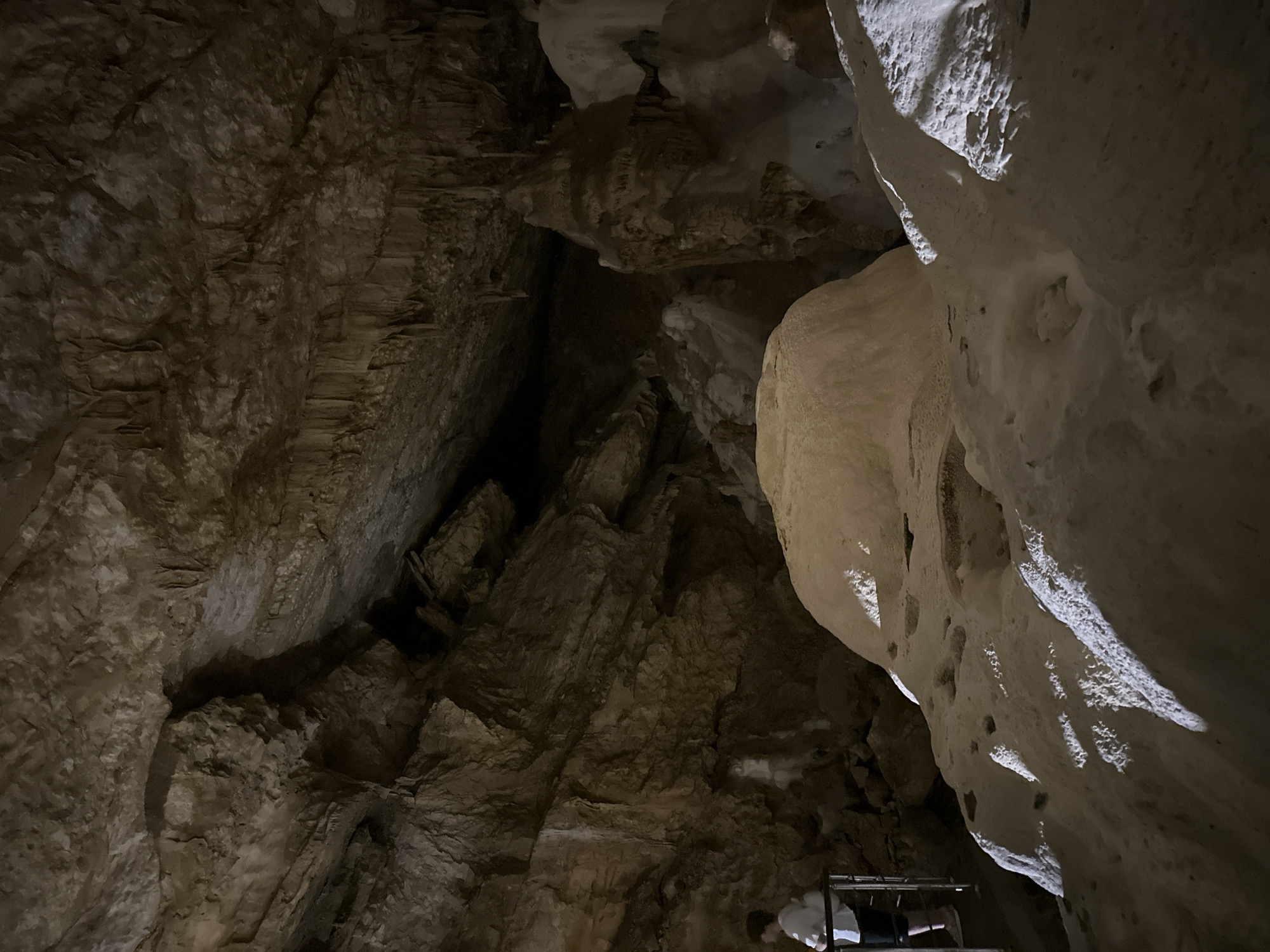 The image shows the interior of a cave. The cave walls and ceiling are rough and textured, with visible stalactites hanging from the ceiling. In the foreground, there are large, smooth, white formations, possibly made of limestone or other mineral deposits. The lighting in the cave is dim, with spotlights highlighting some of the formations. A person in a white shirt and dark shorts is visible on the left side of the image, standing near a railing, suggesting this is a tourist attraction. The person appears to be observing the cave formations. The overall environment is dark, cavernous, and geological. The location is identified as Khostinskiy, Russia.