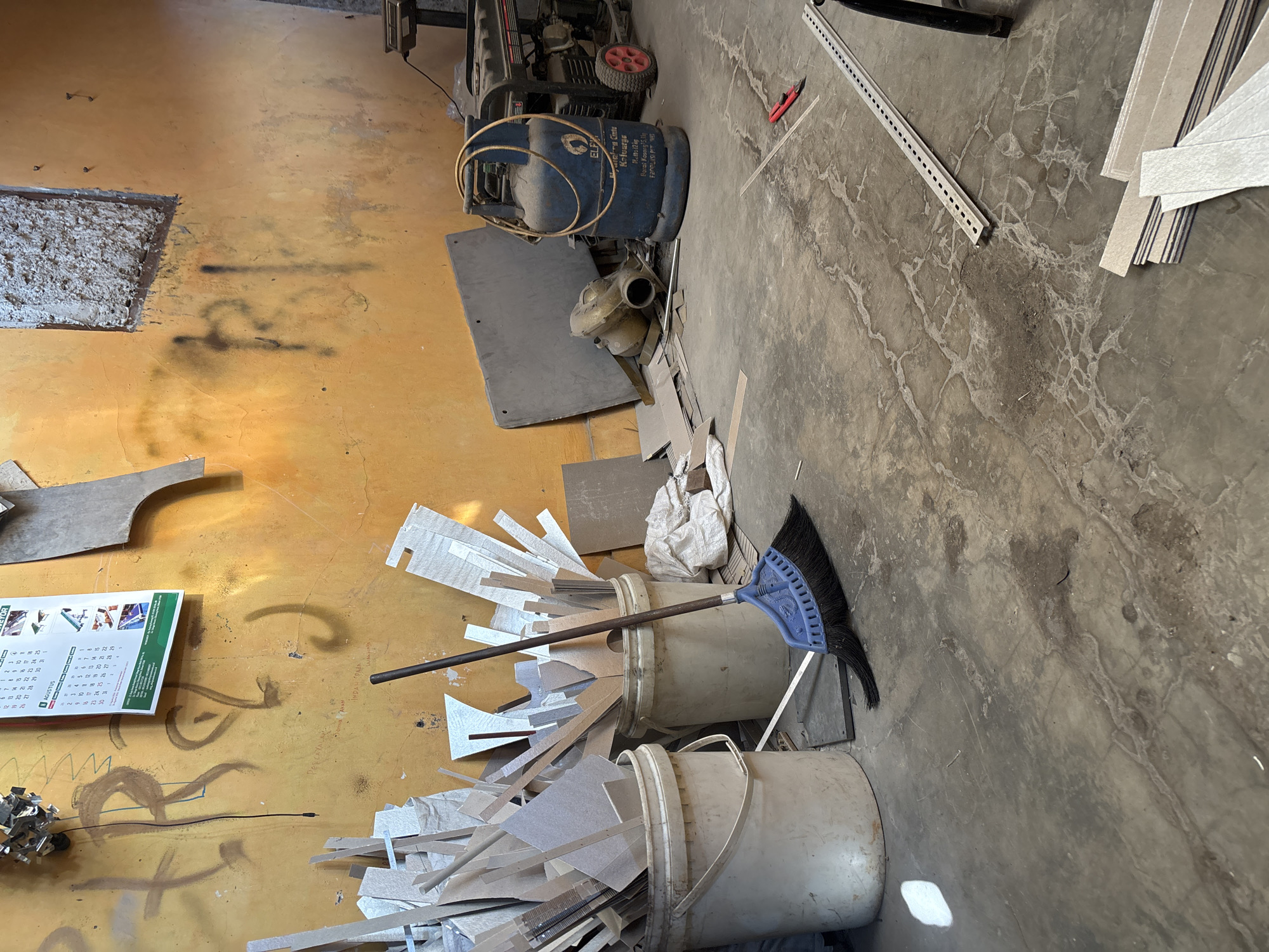 The image depicts an indoor workspace in Telagamurni, Indonesia. The room appears to be cluttered and functional, possibly a workshop or storage area. The walls are painted a bright, somewhat faded yellow and show signs of graffiti. In the foreground, a blue-bristled broom with a dark handle rests on the concrete floor, leaning against two large, off-white plastic buckets. These buckets are filled with various pieces of wood and other materials, likely scraps from construction or crafting.
Towards the right side of the image, a large blue propane tank is visible, connected by a coiled yellow hose. Behind the propane tank sits a portable generator. Scattered on the floor are more pieces of wood, cardboard, and a small red utility knife. In the background, a calendar is hanging on the wall, partially visible. The overall impression is of a work in progress or a space dedicated to a craft or trade.