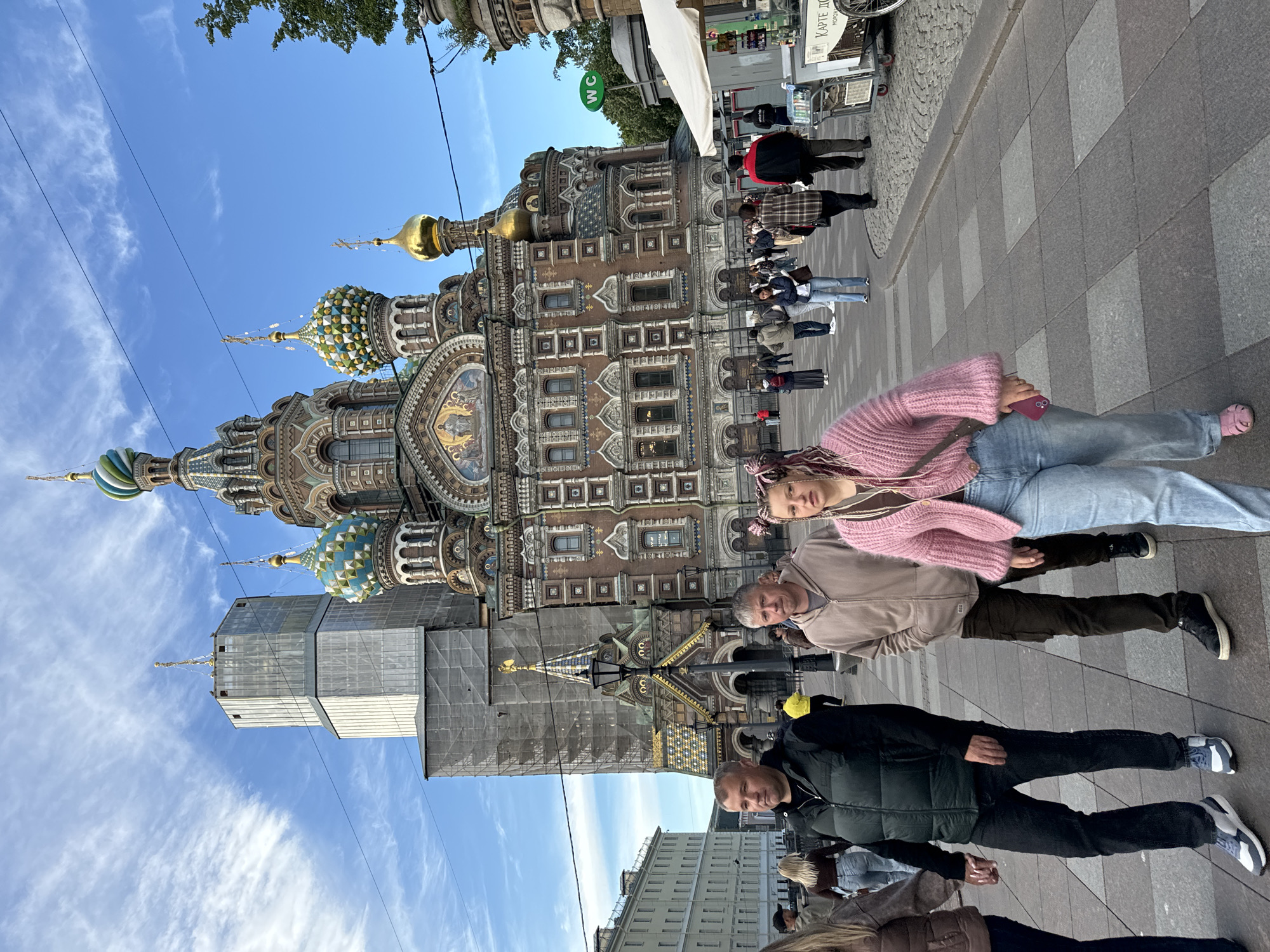 This is a photograph of a group of people posing in front of the Church of the Savior on Spilled Blood in Saint Petersburg, Russia. The church, with its distinct onion domes and ornate facade, dominates the background. The sky is a clear blue with some wispy clouds, suggesting it is daytime, likely with pleasant weather.
In the foreground, four people are lying down or sitting in an unusual pose, almost as if they are resting on the ground. The person closest to the camera is a young woman wearing a pink fuzzy sweater and blue jeans. Next to her is a man in a light brown hooded jacket and dark pants. Another man, wearing a black puffer vest over a black jacket and dark pants, is positioned below them. Partially visible at the bottom is another person. They all appear relaxed and are looking towards the camera.
The sidewalk in front of the church is paved with grey and beige square tiles. To the left of the church, part of a building with scaffolding is visible. In the upper right corner of the image, some market stalls or kiosks can be seen, and a sign with Cyrillic text, possibly advertising "Карте Дом" (Karte Dom), is visible. The overall scene conveys a casual and tourist-like atmosphere, with the people taking advantage of the iconic backdrop for a unique photo opportunity.
