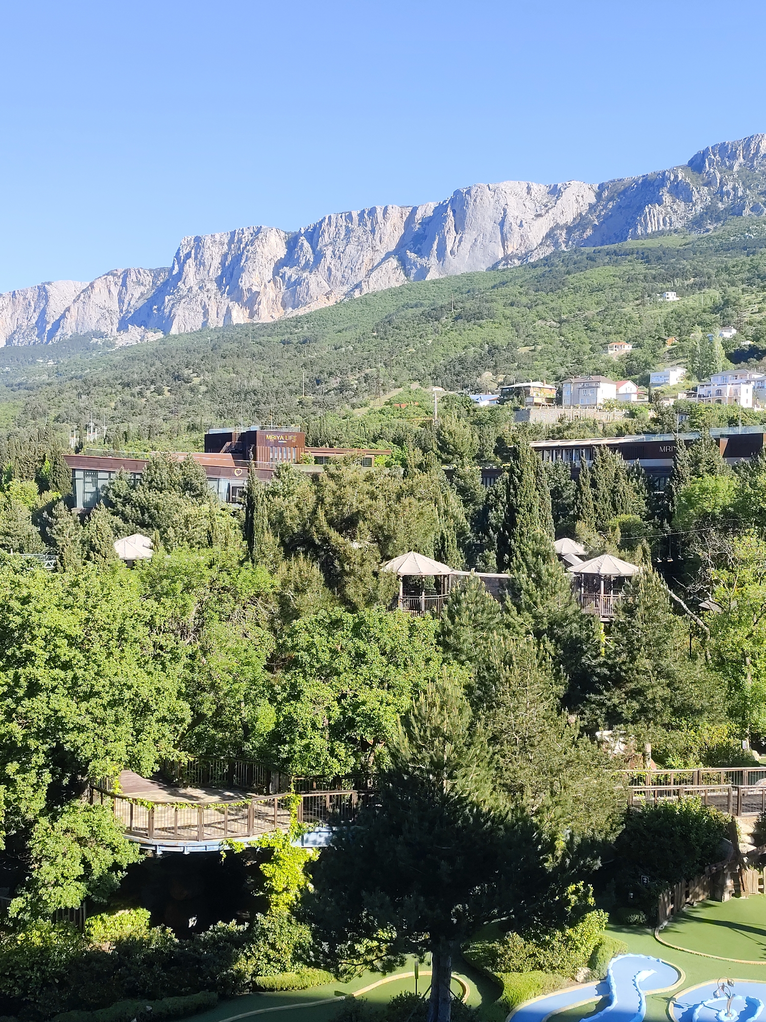 The image shows a mountainous landscape in Симеизский поселковый совет, Ukraine. A long, ridged mountain range dominates the upper portion of the frame. The slopes below are covered with dense green vegetation, including trees and shrubs. Buildings are visible interspersed among the foliage, including a building with "MRIYA LIFE" on it. Gazebos with light-colored canopies are nestled among the trees. In the foreground is a wooden bridge and a miniature golf course. The sky is clear blue.