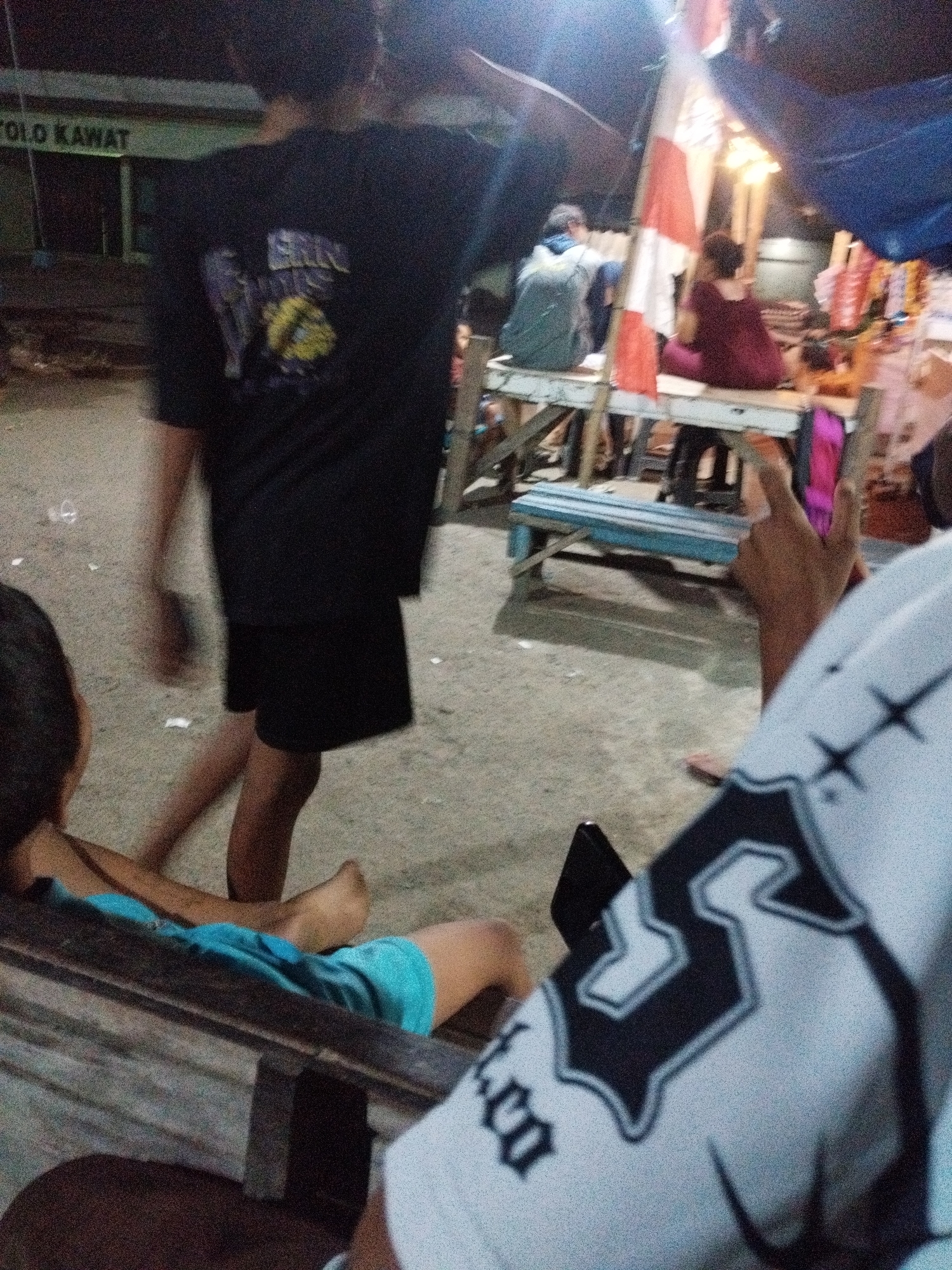 In Kelurahan Cilacap, Kecamatan Cilacap, Indonesia, at night, a group of people are gathered outdoors. A young male in a black t-shirt and black shorts stands in the foreground, with a partial view of his back and legs. He appears to be walking. In the background, to the left, a sign reads "YOLO KAWAT". Several other individuals are seated or standing around a makeshift stall. One person is wearing a grey hoodie and a backpack. Another person, wearing a maroon garment, is seated. A bright red and white flag is displayed. The ground is a dirt or gravel surface, with scattered debris. The overall lighting suggests dusk or nighttime, with artificial light sources illuminating the scene. A person's hand in the lower right corner of the image is making a peace sign, and they are wearing a white garment with black graphic designs.