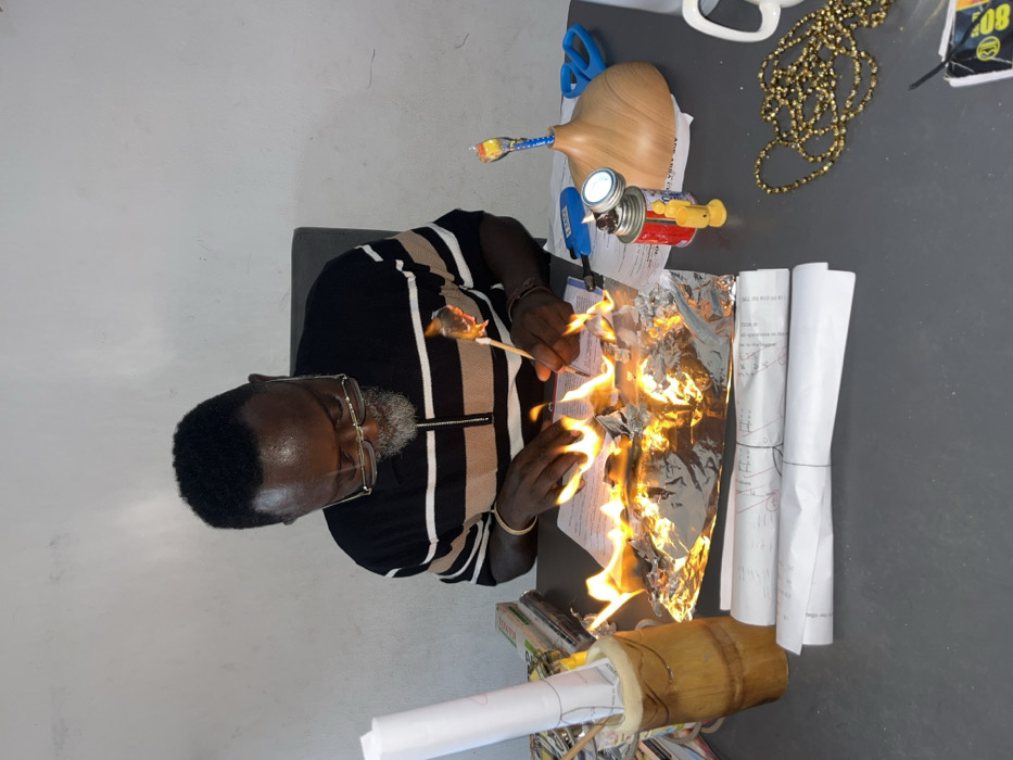 Inside a room in Ado, Nigeria, a middle-aged male with dark skin, short dark hair, a grey beard, and gold-rimmed glasses is seated at a dark grey desk, looking downwards. He wears a black and brown striped polo shirt with a zippered collar.
In front of him, a crumpled piece of aluminum foil is actively burning with orange and yellow flames, resting on a white document. His right hand holds a light-colored stick, the tip of which is also burning, positioned above or within the flames. His left hand is situated near the burning foil.
On the left side of the desk, a bamboo-style cylindrical container holds multiple rolled-up white papers and sticks. Behind this, a stack of books or documents is visible. Two additional rolled-up white papers lie on the desk in front of the bamboo container.
To the right of the central flames, a red and white canister with an attached light or burner stands beside a light-colored, gourd-shaped wooden object. A pair of blue scissors is positioned behind these items. Further to the right, a tangled strand of gold-colored beaded necklace is spread on the desk, next to the handle of a white mug and a small yellow figurine. The background features a plain white wall.