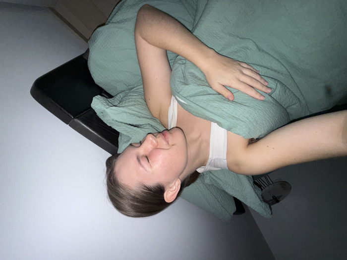 A female individual is depicted lying in a bed in a dimly lit room within the city of Vozdovac Urban Municipality, Serbia. She has light skin, brown hair pulled back, and her eyes are closed, indicating sleep or rest. She wears a white tank top and is partially covered by a textured teal-green duvet and pillow. Her right hand rests upon the duvet over her torso. A dark-colored headboard is positioned behind the pillow. In the upper left background, a ceiling fan with dark blades is partially visible, mounted against a white ceiling. The room features light-colored walls and a sloped ceiling on the right side of the frame.