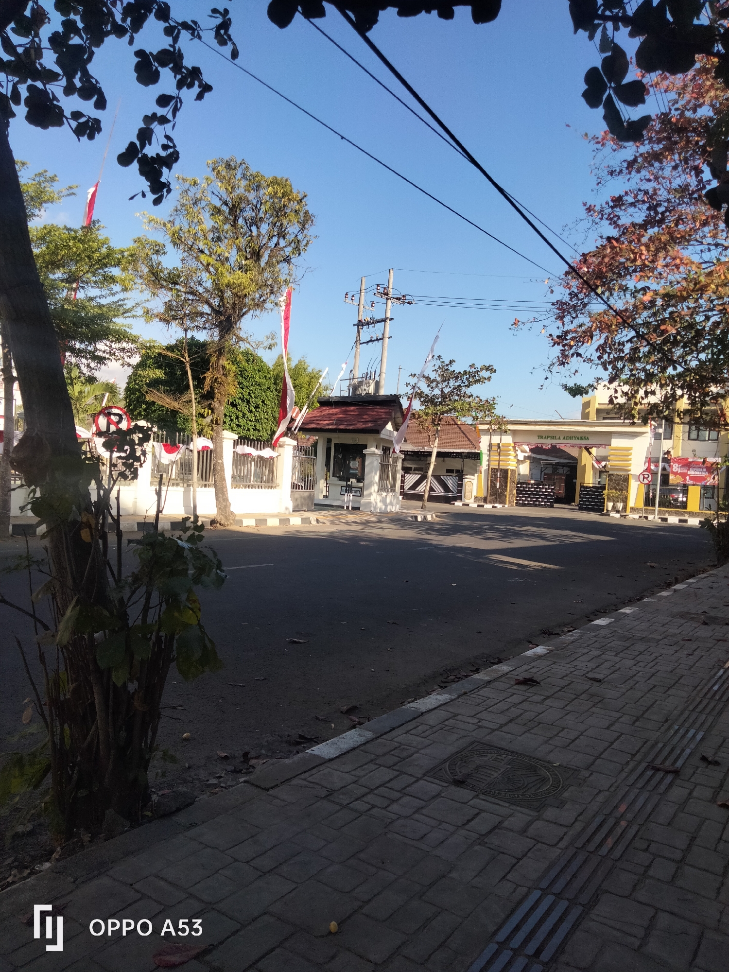 A street scene in Trenggalek, Indonesia, is captured under a clear blue sky. The road is paved and bordered by a sidewalk constructed of grey bricks, featuring tactile paving for the visually impaired. A tree with dark foliage partially obscures the upper left corner of the frame. Red and white Indonesian flags are displayed on poles and strung along a white fence.
In the background, several buildings are visible. The central structure is an archway with the text "TRAPSILA ADHYAKSA" inscribed above it. This building has a yellow and white facade, with decorative black and yellow columns flanking the entrance. To the left, another building with a red roof and white walls is partially visible. A "no parking" sign is prominent on the left side of the street. Trees with green and brown foliage are scattered throughout the scene. The overall impression is one of a well-maintained public area, possibly near a government or official building, decorated for a national celebration. The lighting suggests it is daytime, likely morning or late afternoon due to the warm tones and distinct shadows. The bottom of the image includes a watermark indicating it was taken with an OPPO A53 phone.